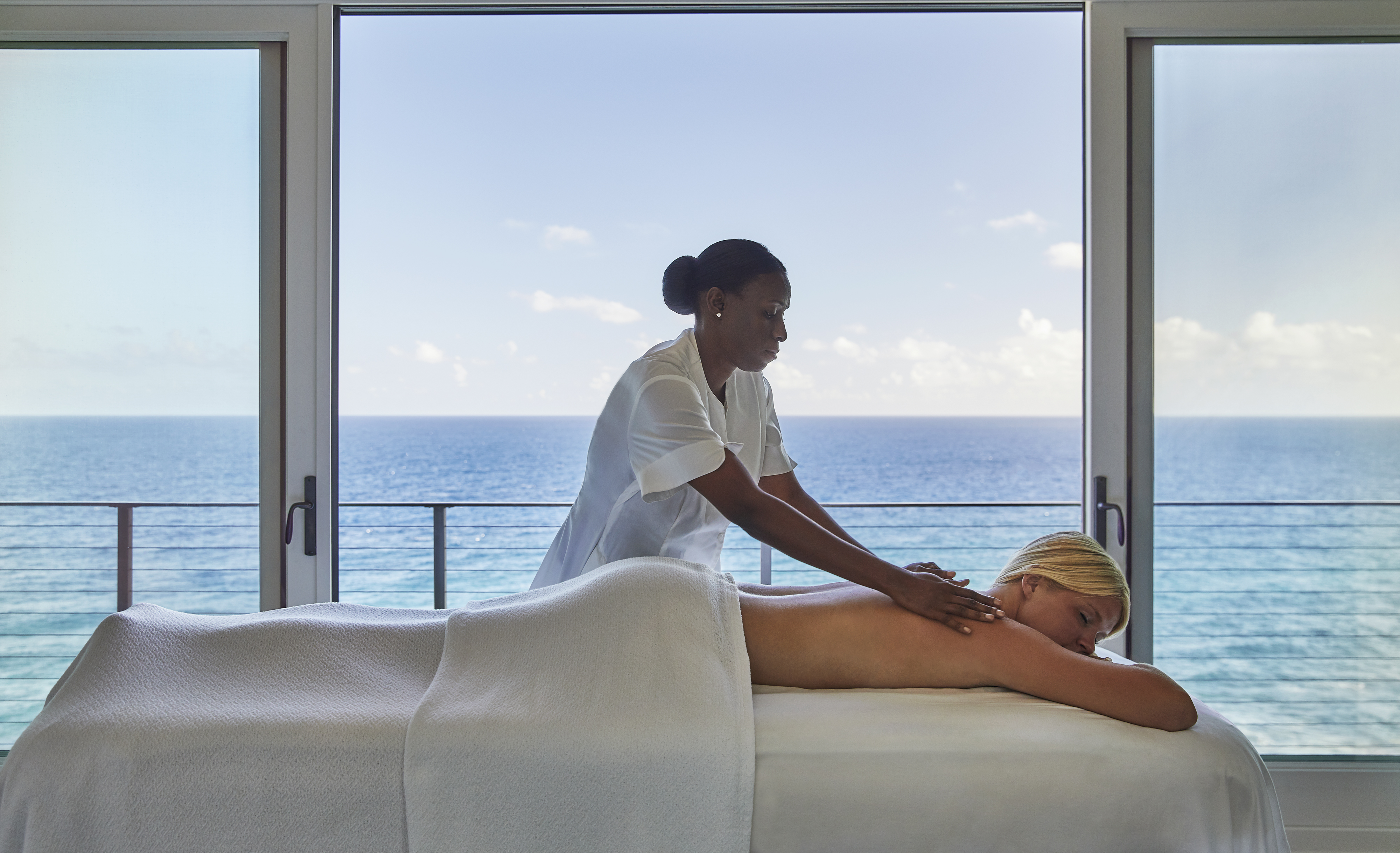 A massage therapist from Oil Nut Bay Spa gives a back massage to a person lying on a massage table covered with a white towel. The room has large windows overlooking the ocean, with a clear, blue sky in the background, creating a serene and relaxing atmosphere.