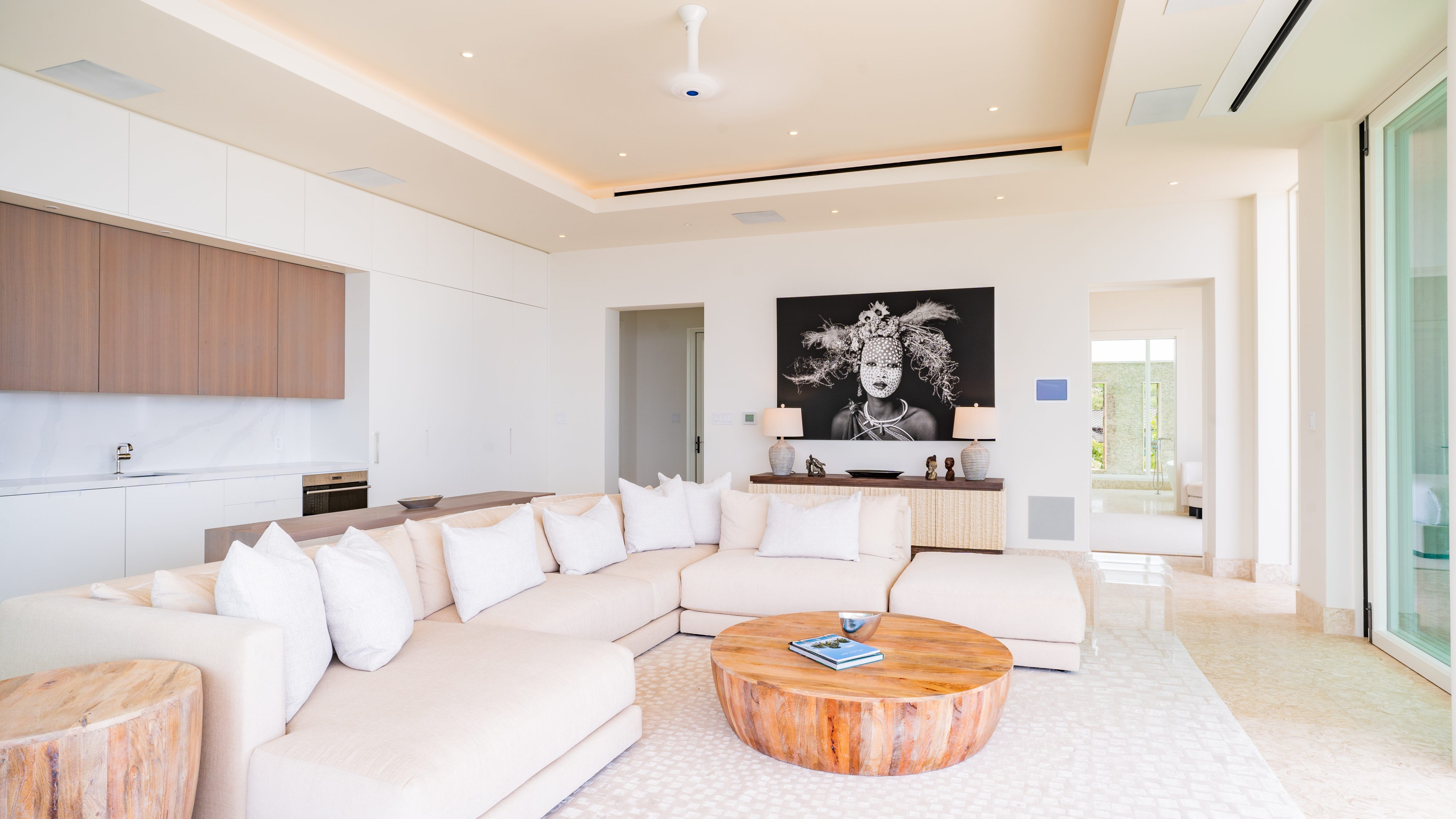 A modern villa living room with a large beige sectional sofa adorned with white pillows. A round wooden coffee table sits in front. The walls are white, featuring a black-and-white portrait. Recessed lighting and large windows provide natural light. A kitchenette is visible.