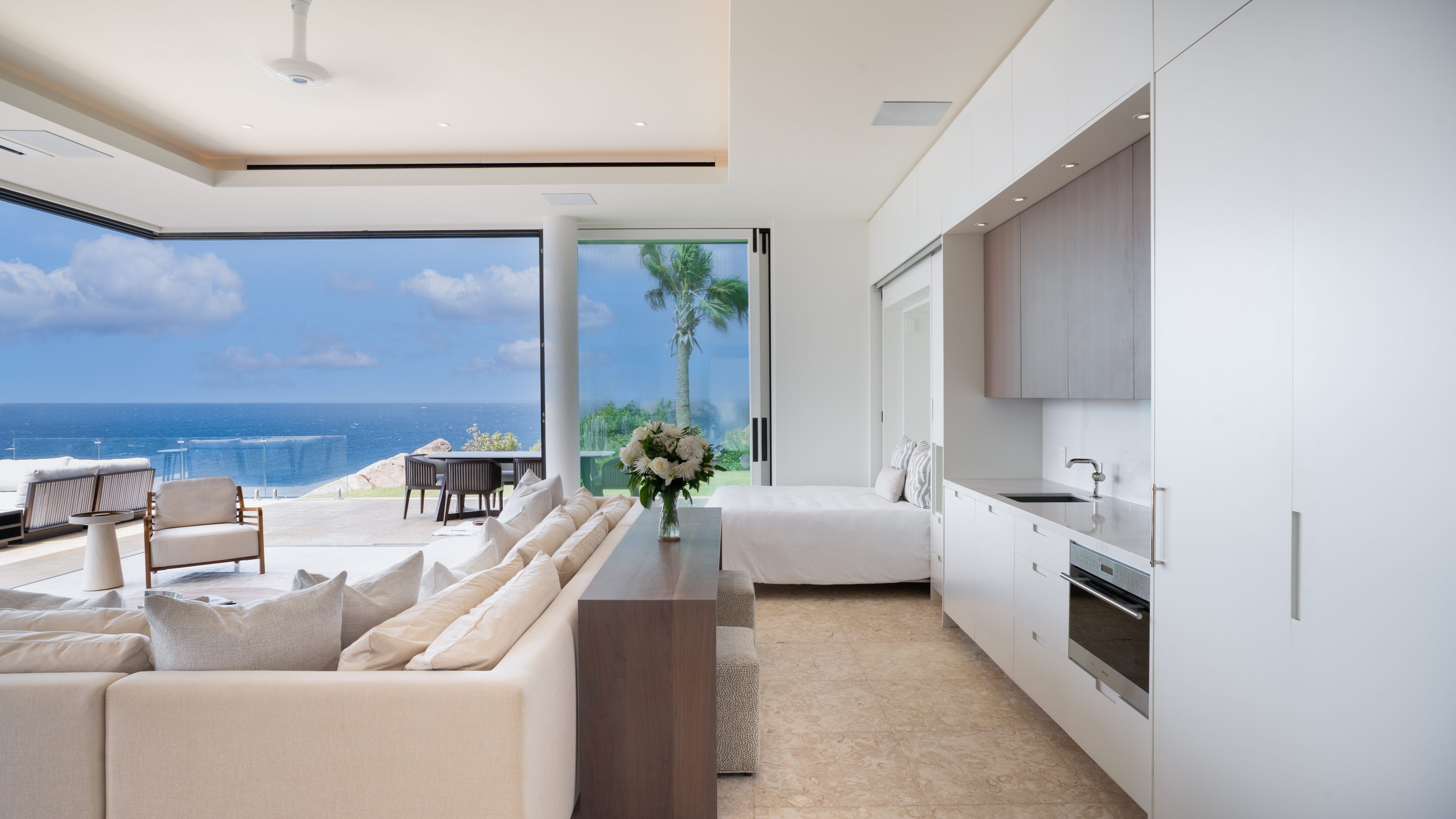 A spacious, modern living area with white walls and light-colored furniture overlooks a breathtaking ocean view at Starlight Villa. Large floor-to-ceiling windows and sliding doors open up to an outdoor patio with seating and a pool, while a small kitchenette is visible on the right.