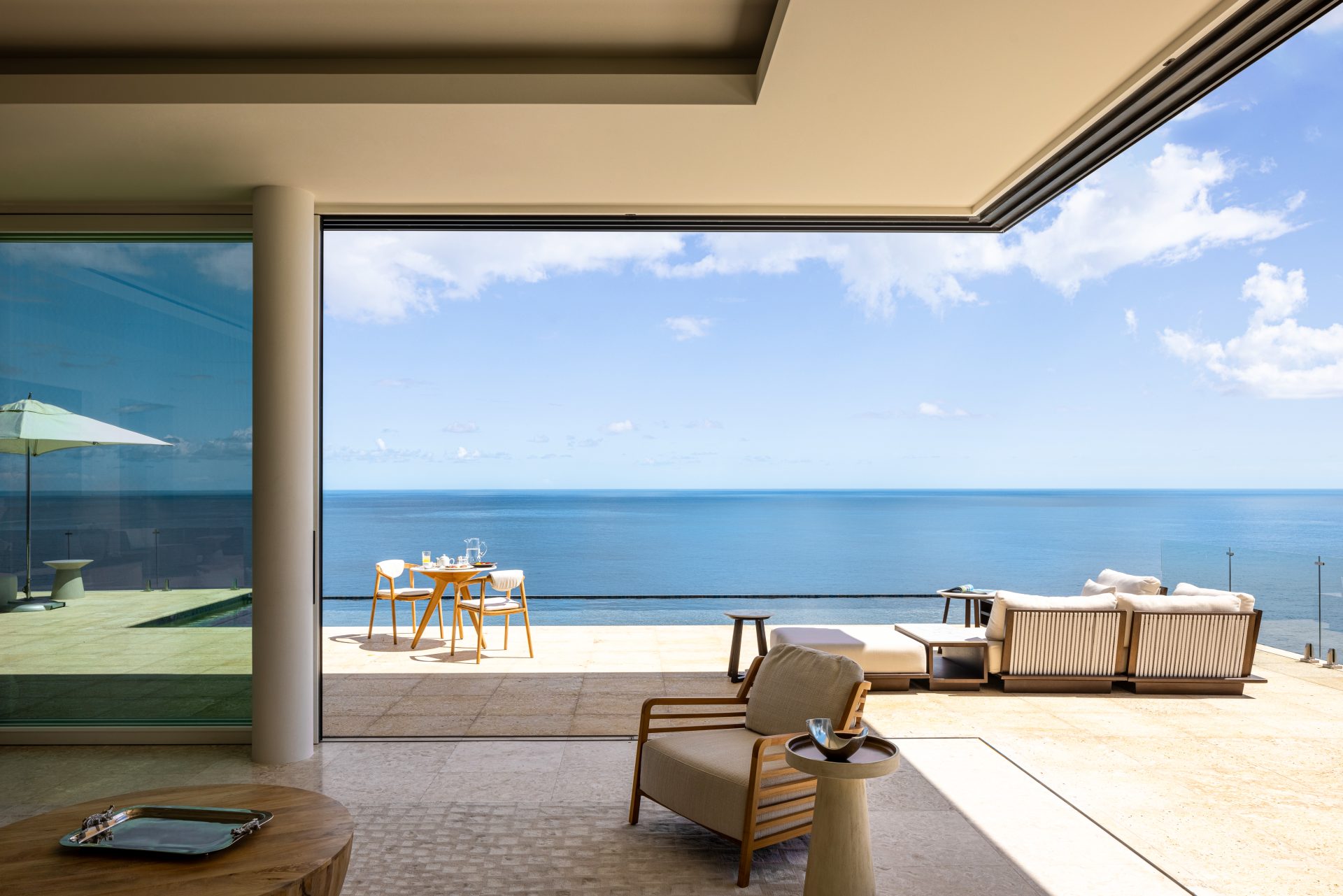 A modern open-concept living space with a view of the ocean at Villa Starlight. Outdoor seating includes a table with chairs, a cushioned lounge area, and a wooden chair with a small table. The space is bathed in natural light, with a clear blue sky and calm sea in the background.