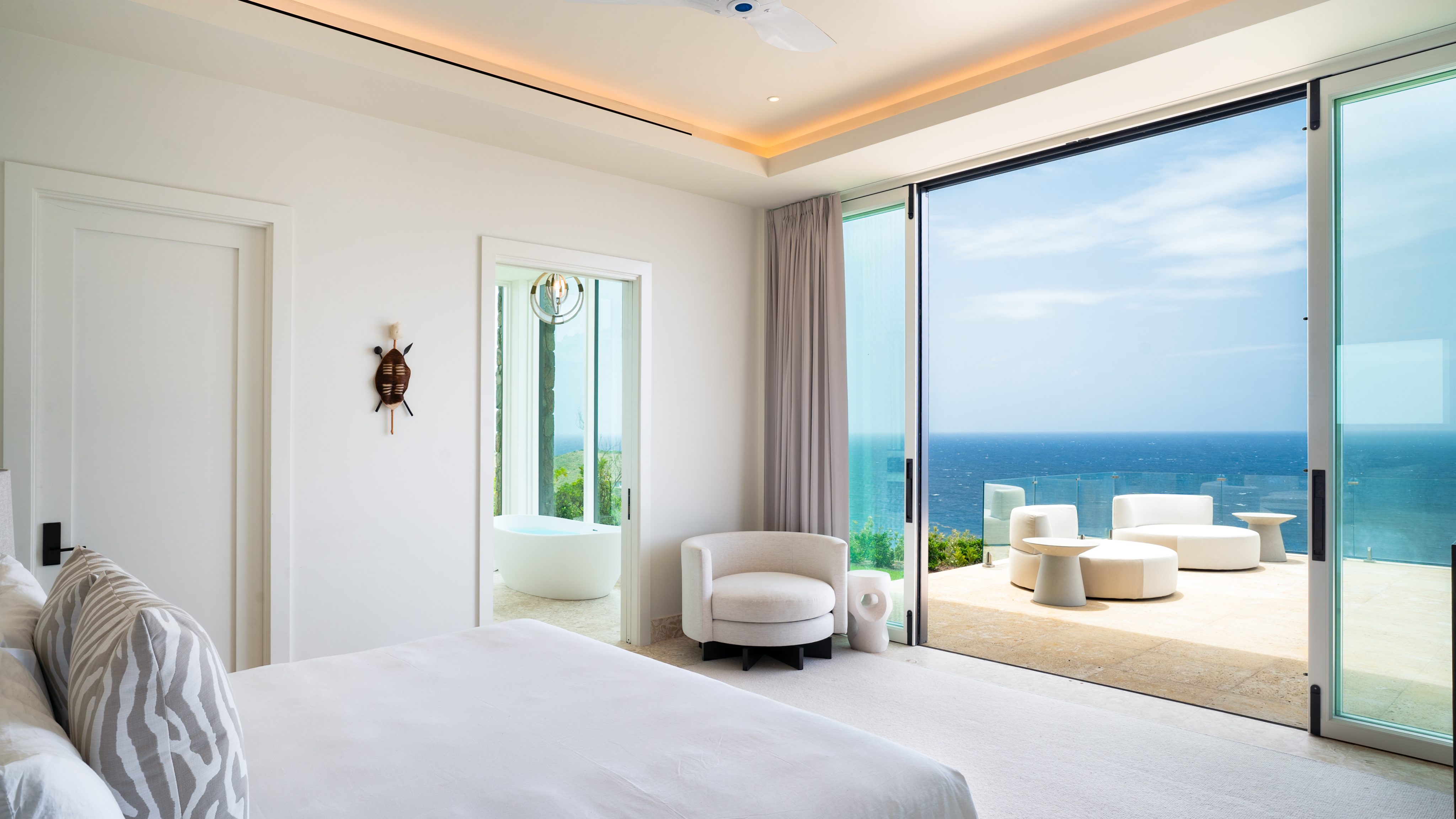 Starlight Villa offers a modern, minimalist bedroom featuring a large bed with white linens, an armchair, and an open door to a bathroom with a freestanding bathtub. Floor-to-ceiling windows provide stunning ocean views, while the outdoor patio is furnished with elegant white furniture.