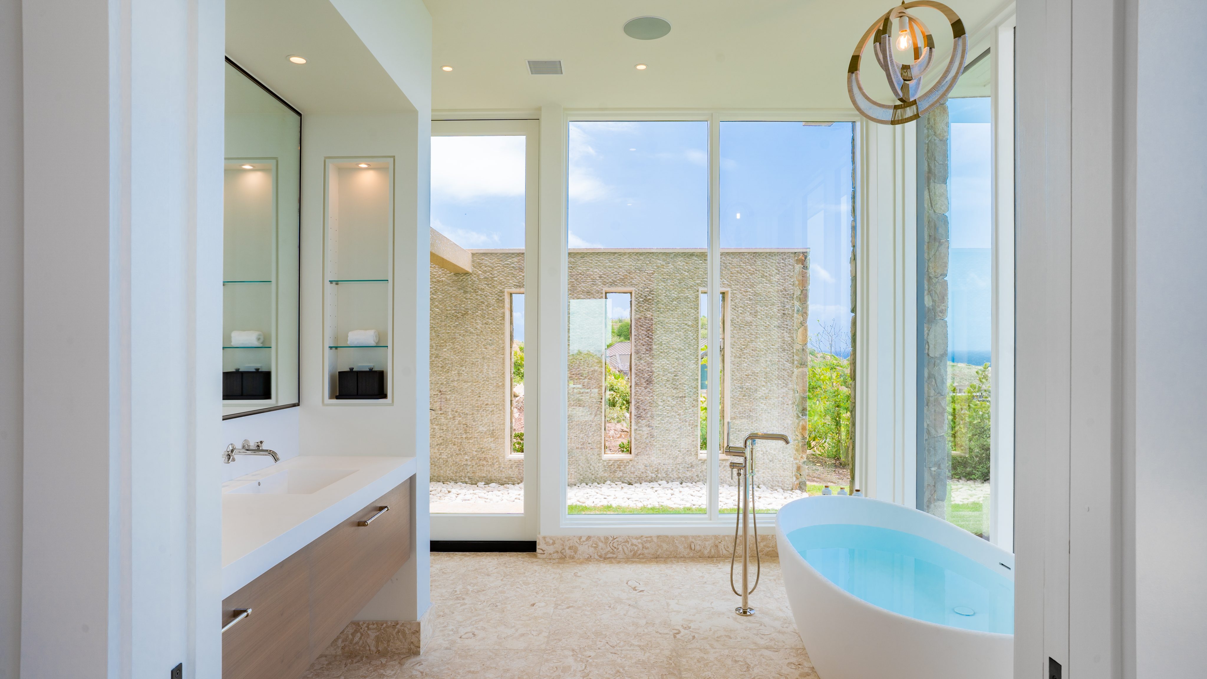 A modern bathroom at Starlight Villa features a large standalone bathtub positioned next to floor-to-ceiling windows with a scenic view. The room includes a wide vanity with a sink and mirror on the left, minimalist decor, and a sleek light fixture hanging from the ceiling.