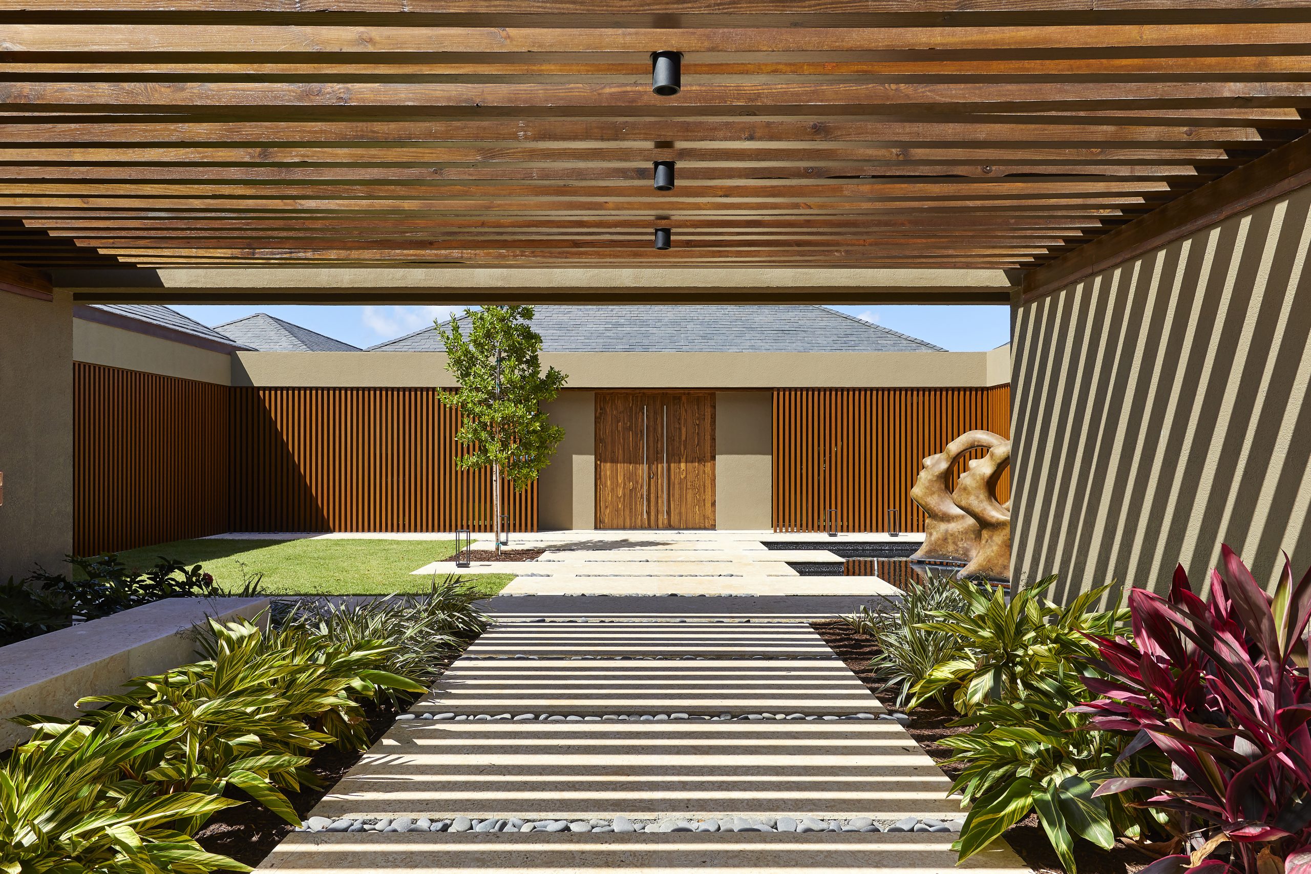 An outdoor passageway with wooden slats forming a pergola overhead leads to a courtyard featuring a modern building with wooden doors. Decorative bushes and plants line the pathway, and a stone sculpture is visible near a small pond, creating an inviting atmosphere for this beachfront property.