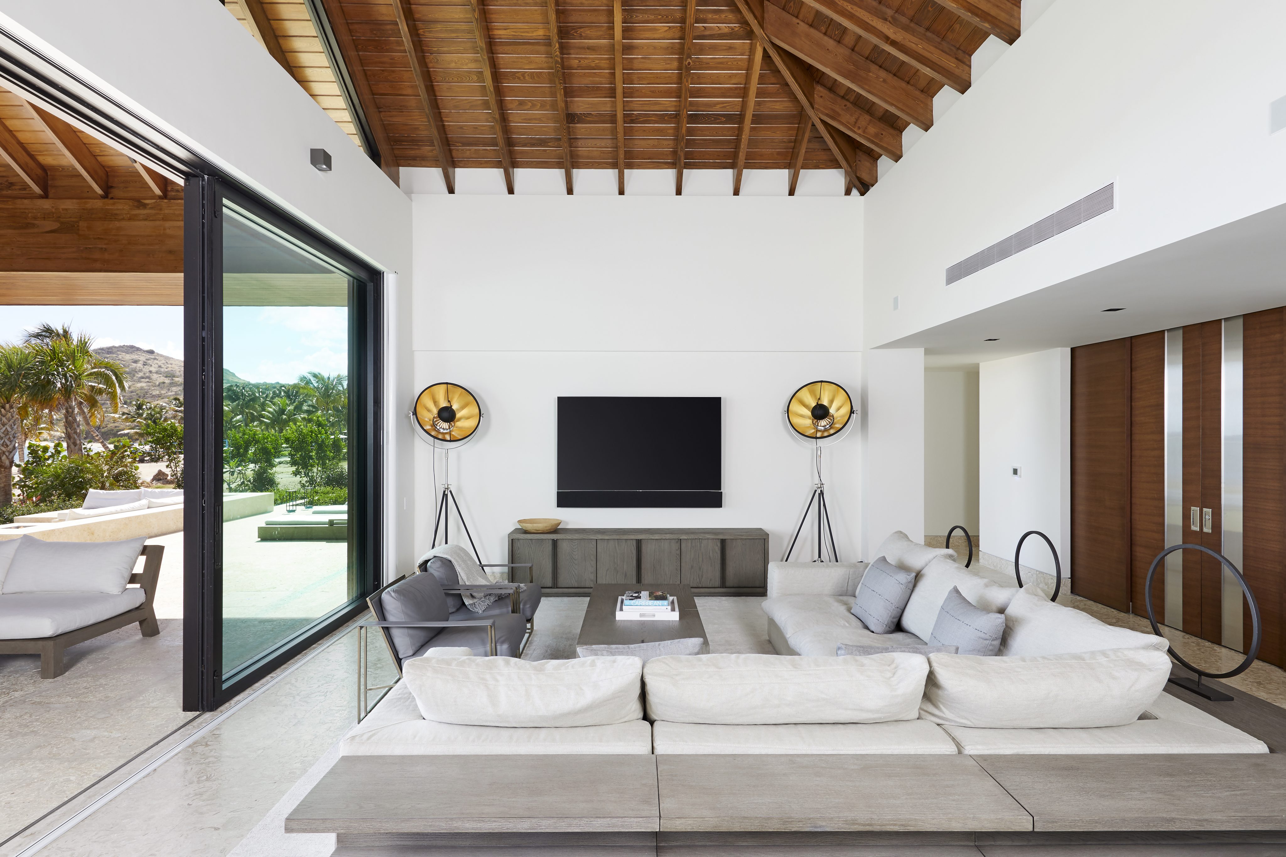 Modern living room with a high wooden ceiling and large sliding glass doors leading to an outdoor patio, perfect for a coastal vacation rental. Features a wall-mounted TV, gray sectional sofa, contemporary floor lamps, and neutral-toned furniture. Bright and spacious with a view of greenery outside.