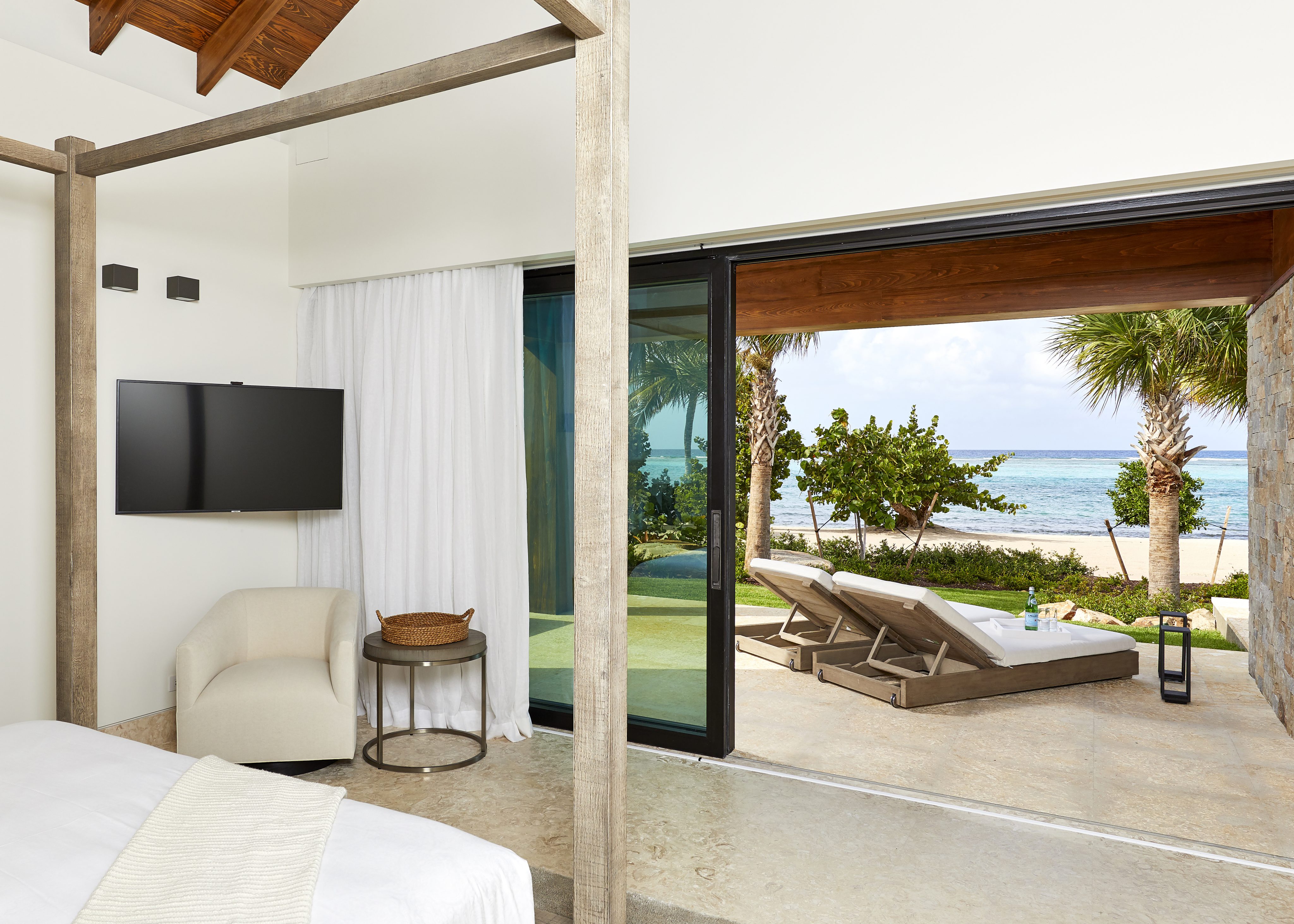 A luxurious bedroom in this seaside accommodation features a canopy bed, a wall-mounted TV, and a cozy armchair. Sliding glass doors open to a patio with lounge chairs overlooking the sandy beach and ocean, framed by palm trees and lush foliage—perfect for your next vacation rental.