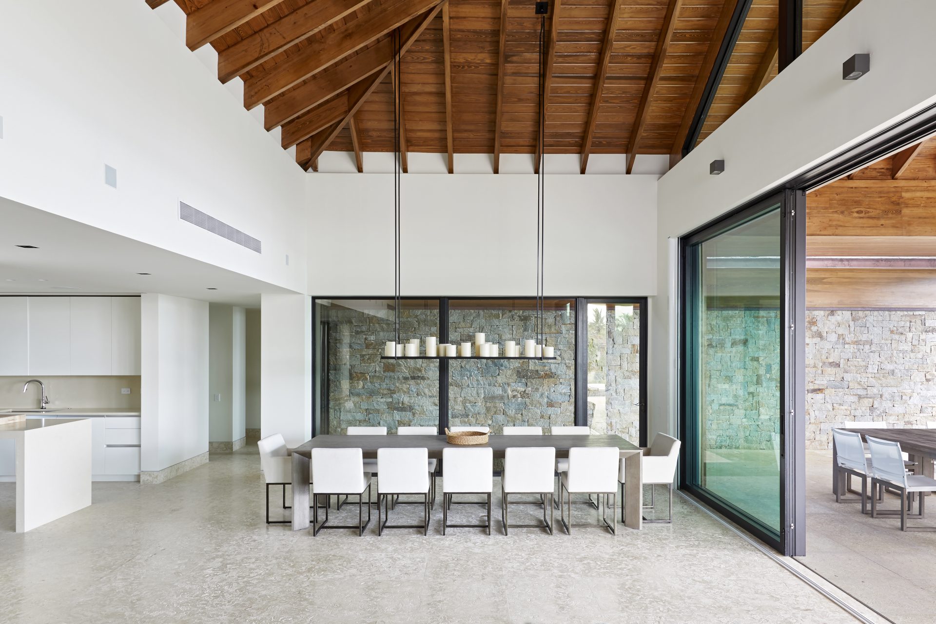 Spacious modern dining area with a large rectangular table and white chairs. The room features high wooden ceilings, a stone wall, and large sliding glass doors opening to an outdoor patio. Adjacent is a sleek, minimalist kitchen with a white island and cabinetry, embodying the essence of a beachfront property.