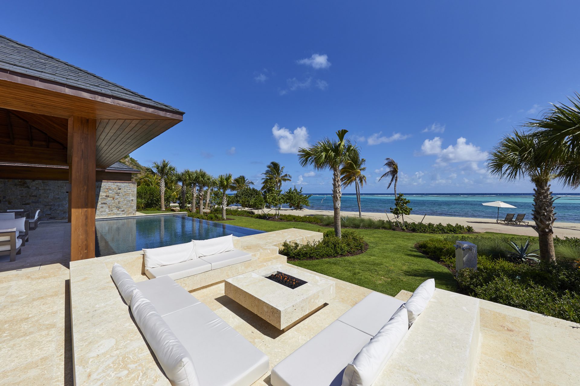 A luxurious oceanfront patio with white cushioned seating around a fire pit, overlooking a spacious lawn, palm trees, and the beach. This stunning vacation home features a modern house with stone accents and a pool to the left. The clear blue sky completes this idyllic seaside rental.