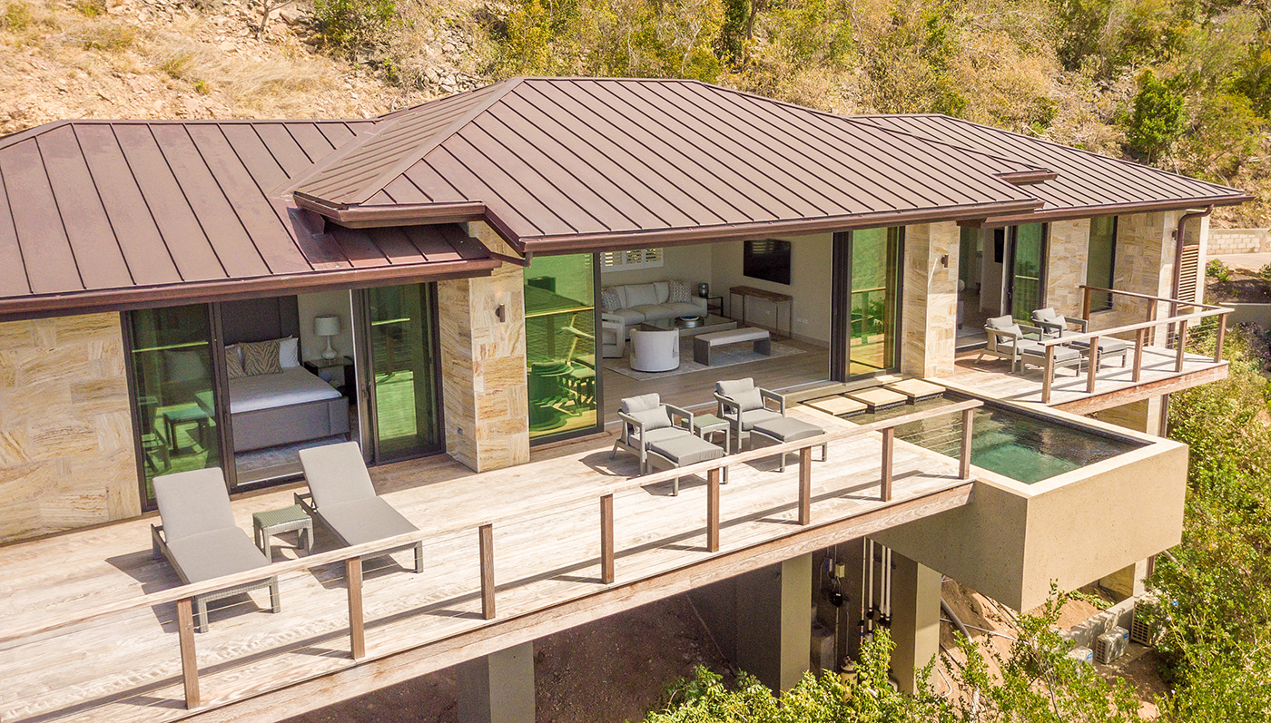 A luxurious vacation rental, Sur Villa features a spacious deck with lounge chairs, an outdoor seating area, and a private pool. The modern structure has large glass doors, a metal roof, and is surrounded by lush greenery and a hillside landscape.