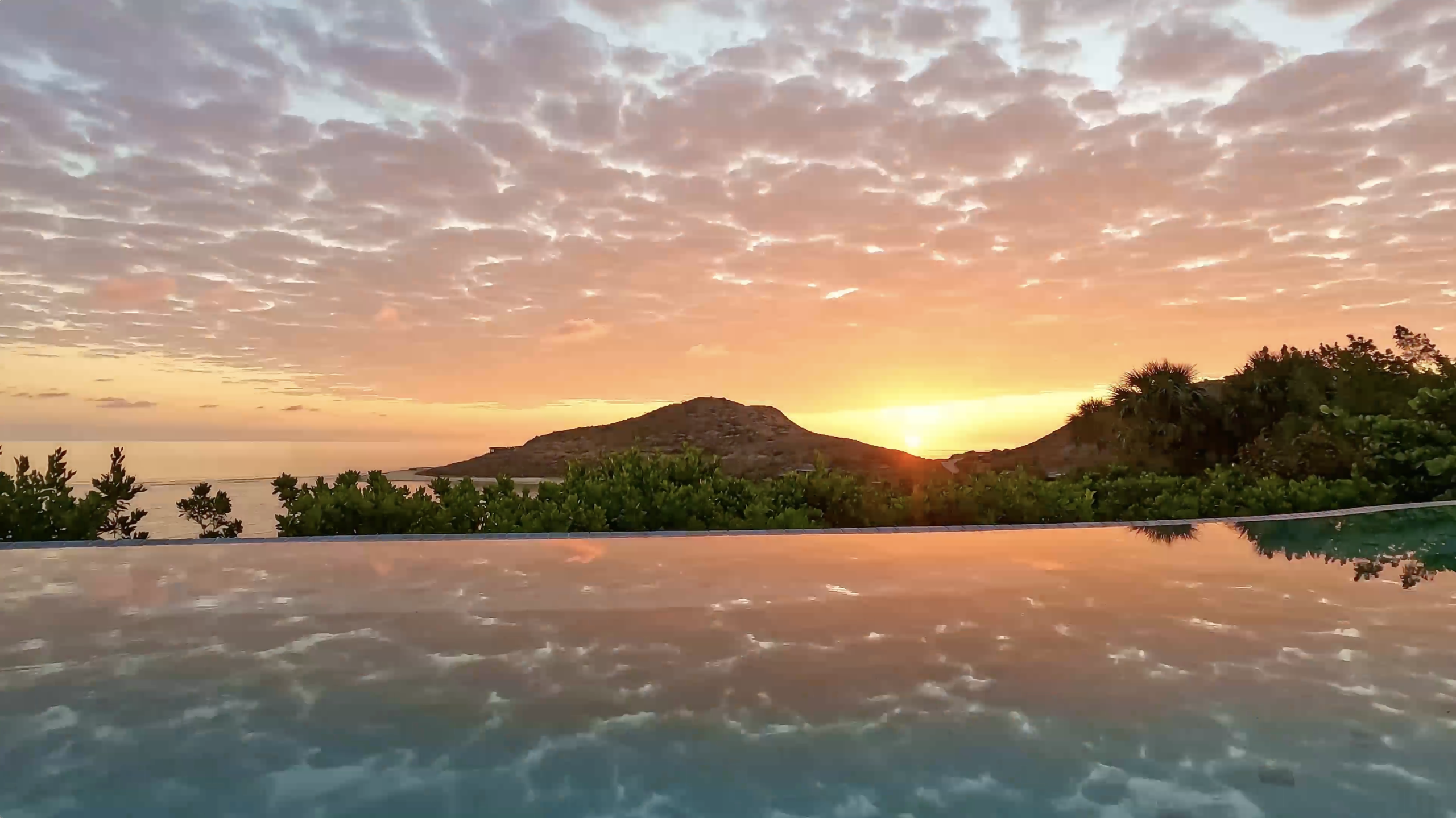 A tranquil scene of a sunset with a vibrant sky filled with clouds reflecting on a calm body of water. In the background, the silhouette of a small hill and lush greenery adds to the serene beauty, reminiscent of an idyllic villa lifestyle.