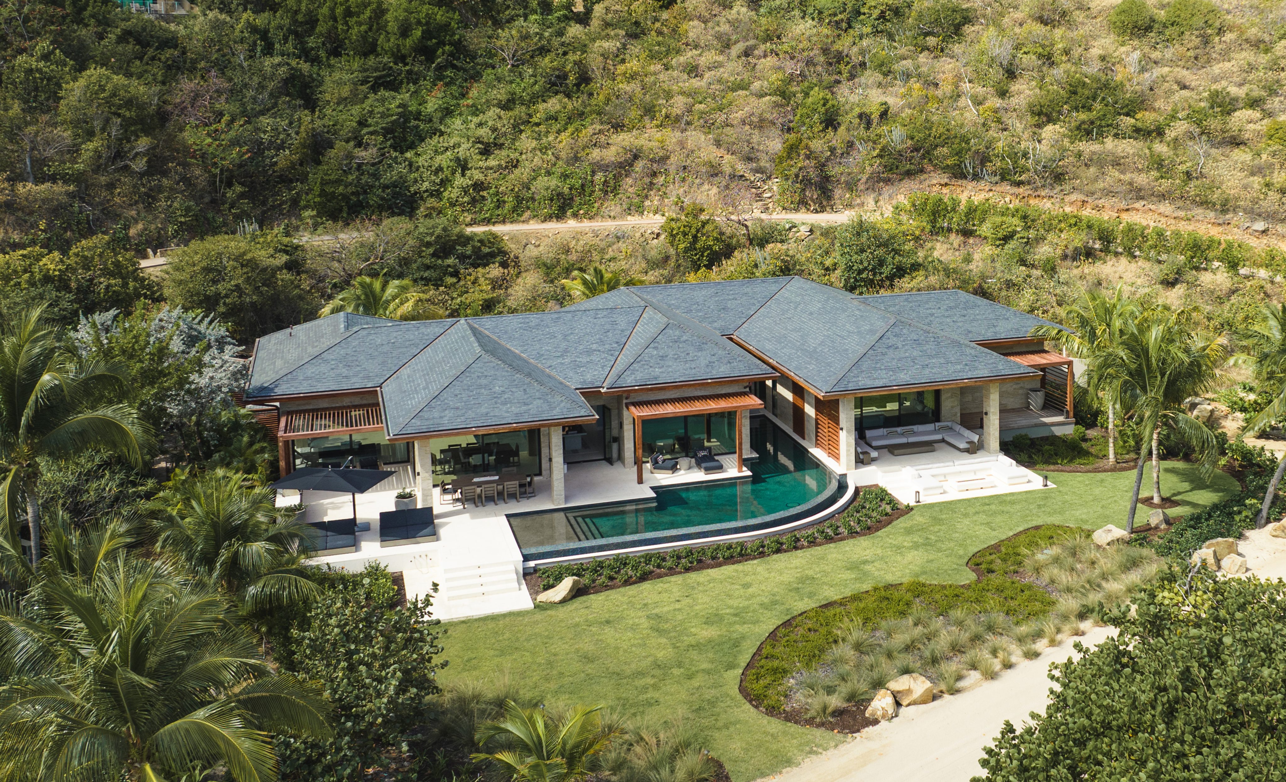 Aerial view of a luxurious modern villa with a blue tile roof, nestled in a lush, tropical landscape. The property, part of the exclusive Oil Nut Bay Rentals, features a large central swimming pool surrounded by manicured lawns, multiple outdoor seating areas, and a pathway leading to the entrance.