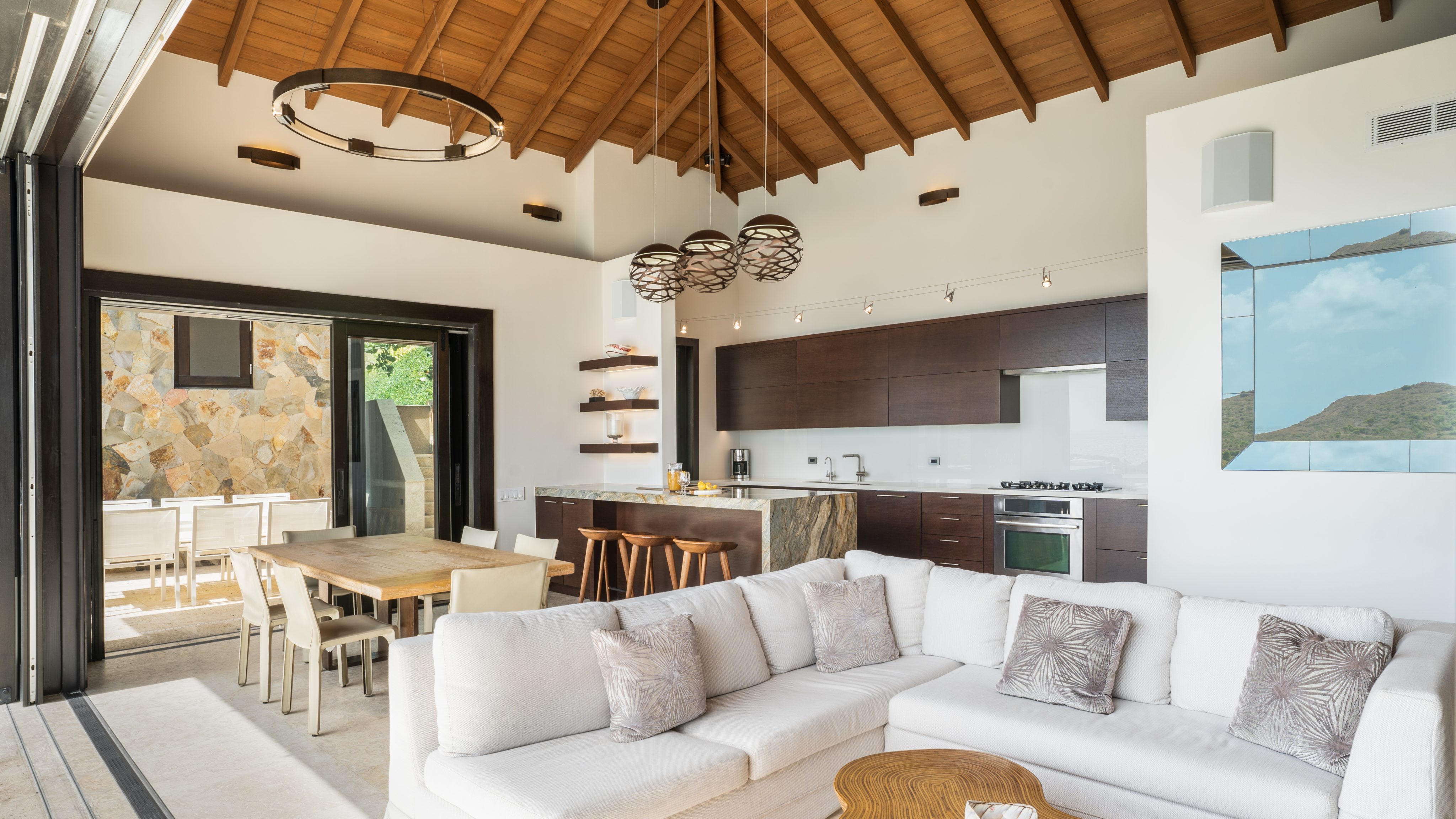 A modern open-concept living space in a stunning villa at the water's edge features a white sectional sofa with patterned pillows, a wooden dining table, and a sleek kitchen with dark cabinets and bar stools. The room has a high wooden ceiling, pendant lighting, and large sliding doors opening to an outdoor area.