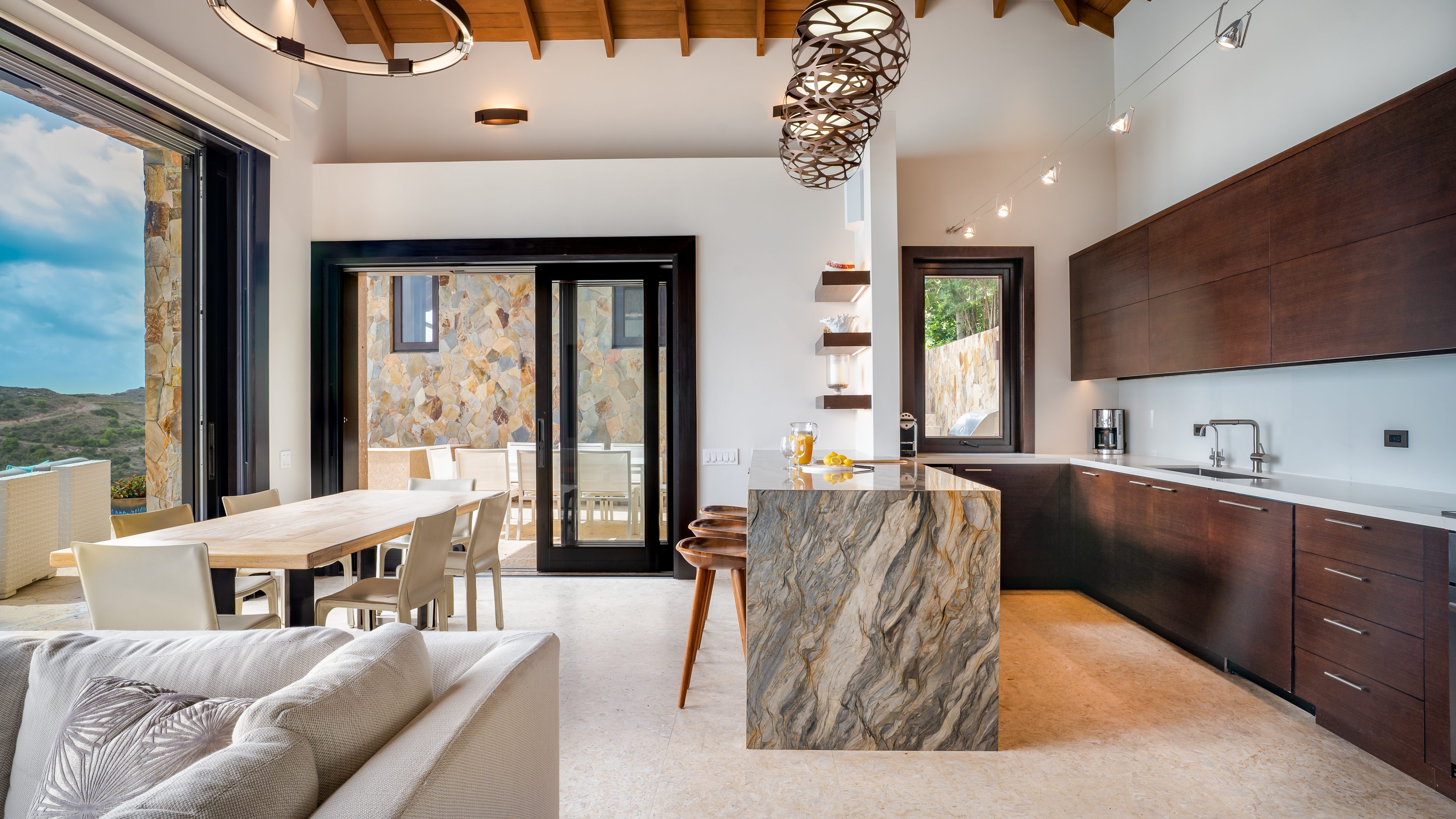 A modern kitchen and dining area with high wooden ceilings, large windows, and stone countertops. The space includes a dining table with chairs, a kitchen island with stools, and contemporary light fixtures. Through the sliding doors is the patio of this elegant Villa at Water's Edge, featuring serene outdoor seating.