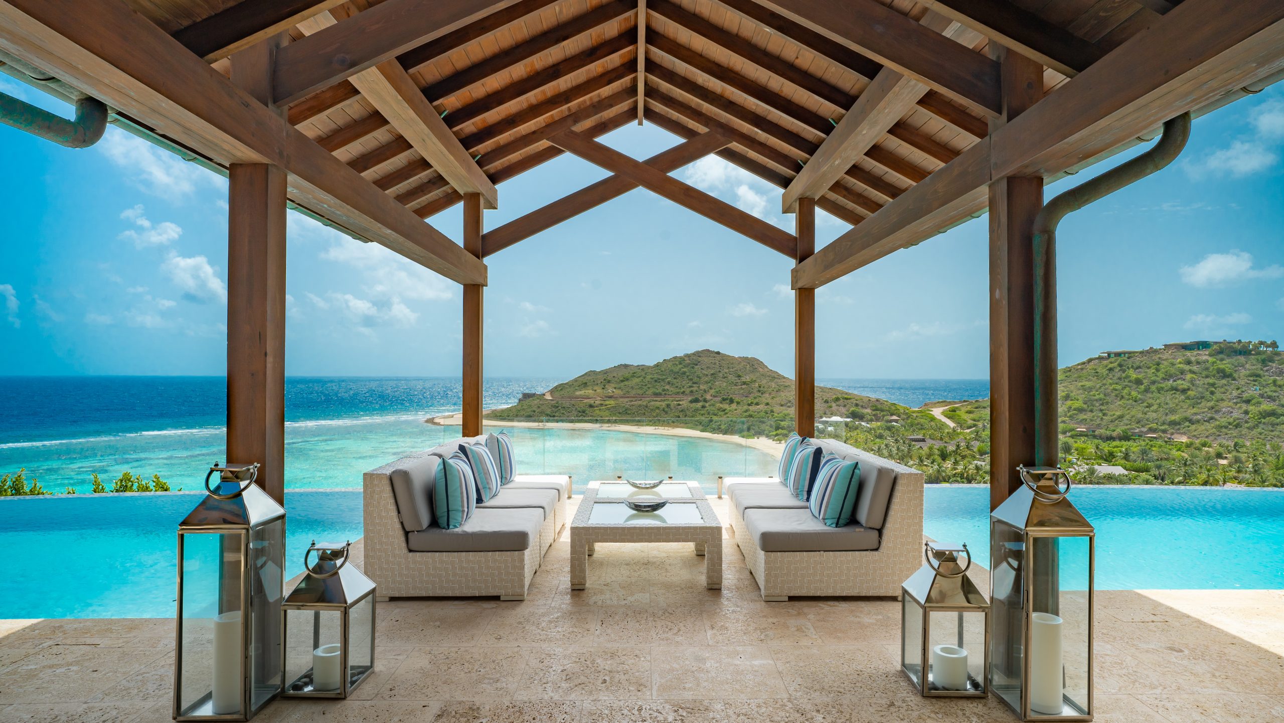 A stunning outdoor lounge area at the water's edge features a covered wooden pergola with white wicker sofas and a coffee table, overlooking an infinity pool and a serene tropical landscape with ocean views and a distant island. Two lantern-style lights adorn the villa patio.