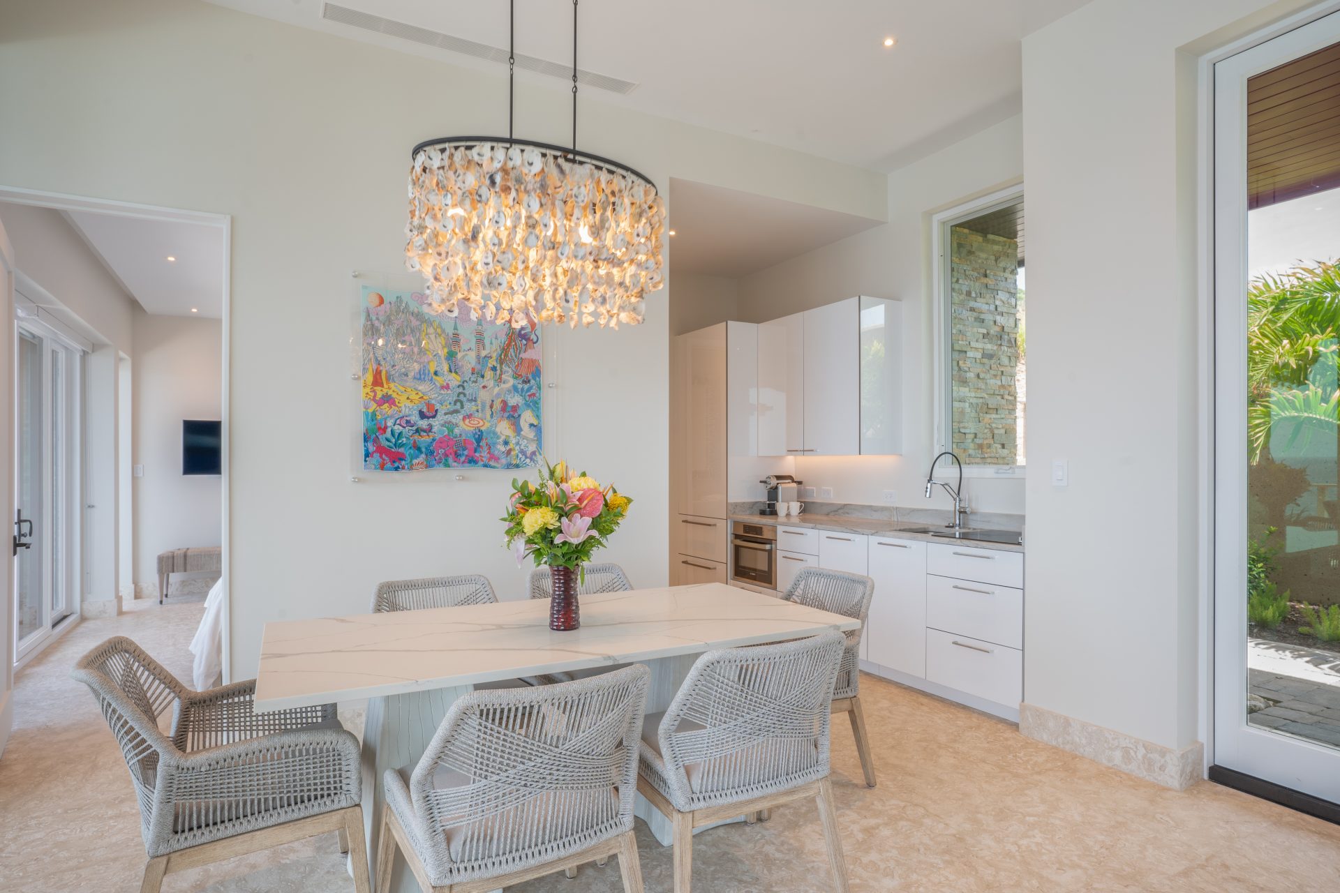 A modern dining area in Whale Point Villa features a white marble table surrounded by six wicker chairs. A vase of colorful flowers centers the table, beneath a decorative chandelier. The background showcases a white kitchen with cabinets, a large colorful painting, and a doorway leading outside.