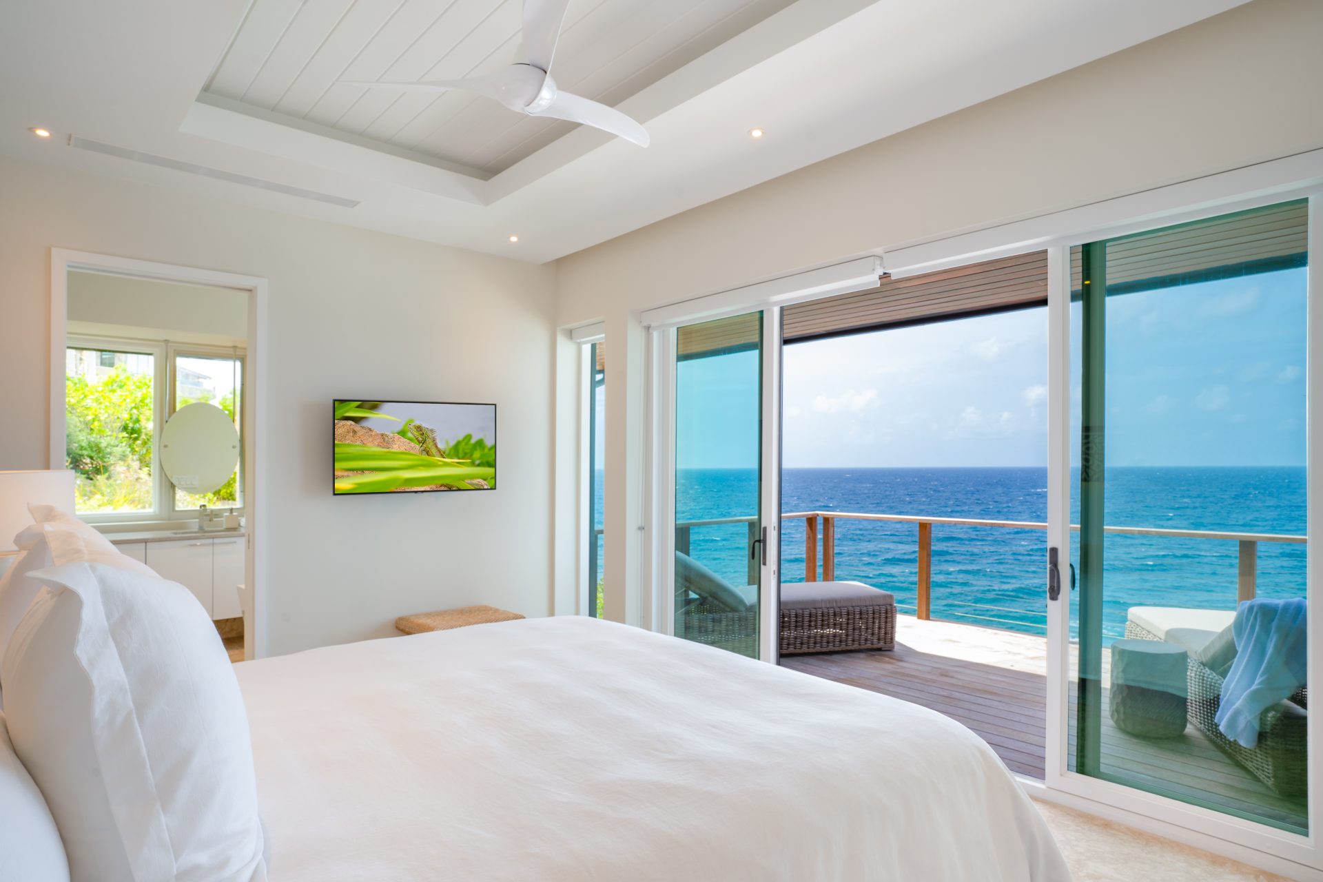 A bright, modern bedroom at Whale Point Villa features white bedding, a ceiling fan, and a large sliding glass door leading to a balcony with a stunning ocean view. A flat-screen TV is mounted on the wall, and there's a vanity area with a round mirror near a window showcasing lush greenery.