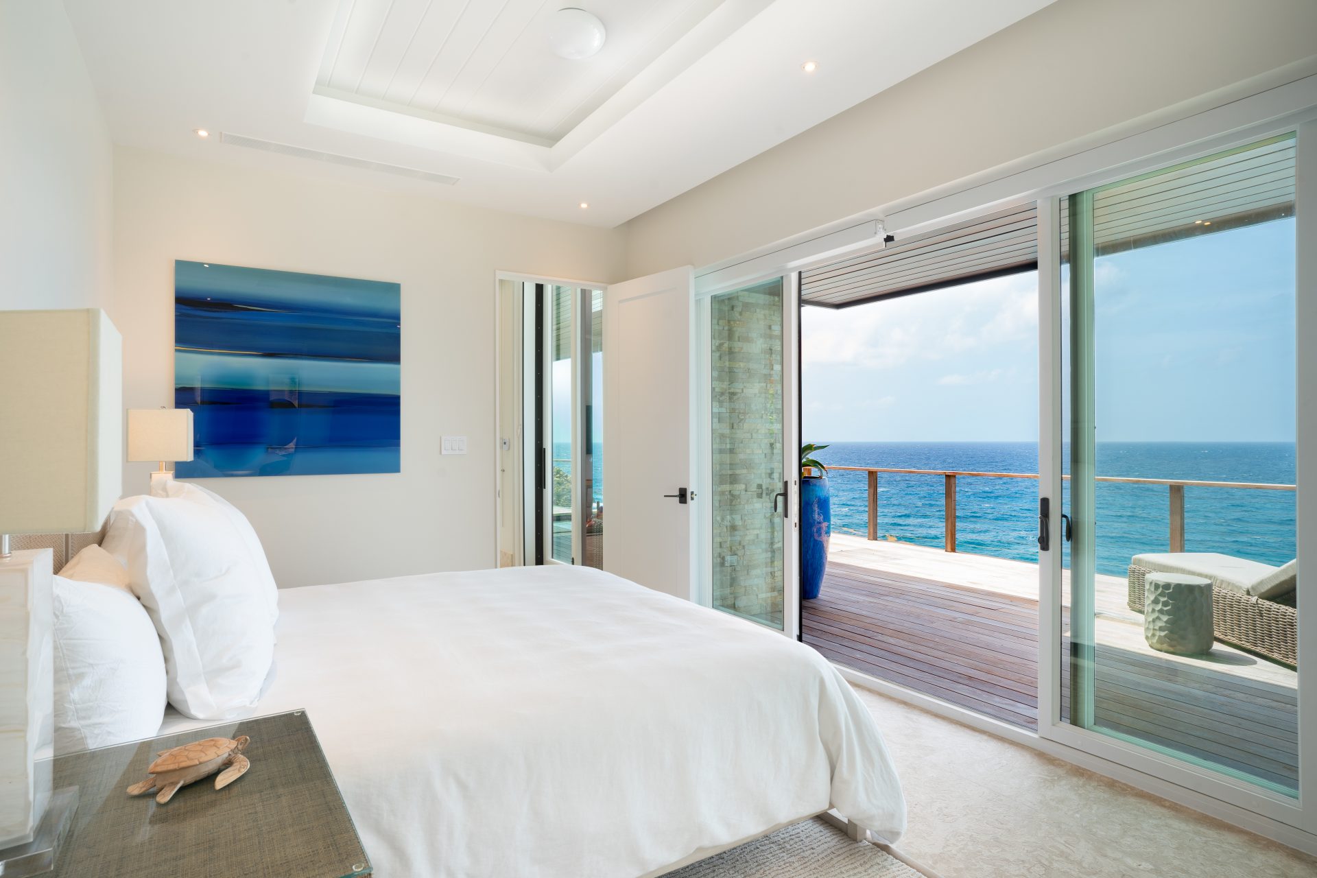 A bright and modern bedroom at Whale Point Villa features a neatly made white bed, a nightstand holding a lamp and decorative items, and a large painting on the wall. Glass sliding doors open to a wooden deck with ocean views, showcasing patio furniture and a potted plant.