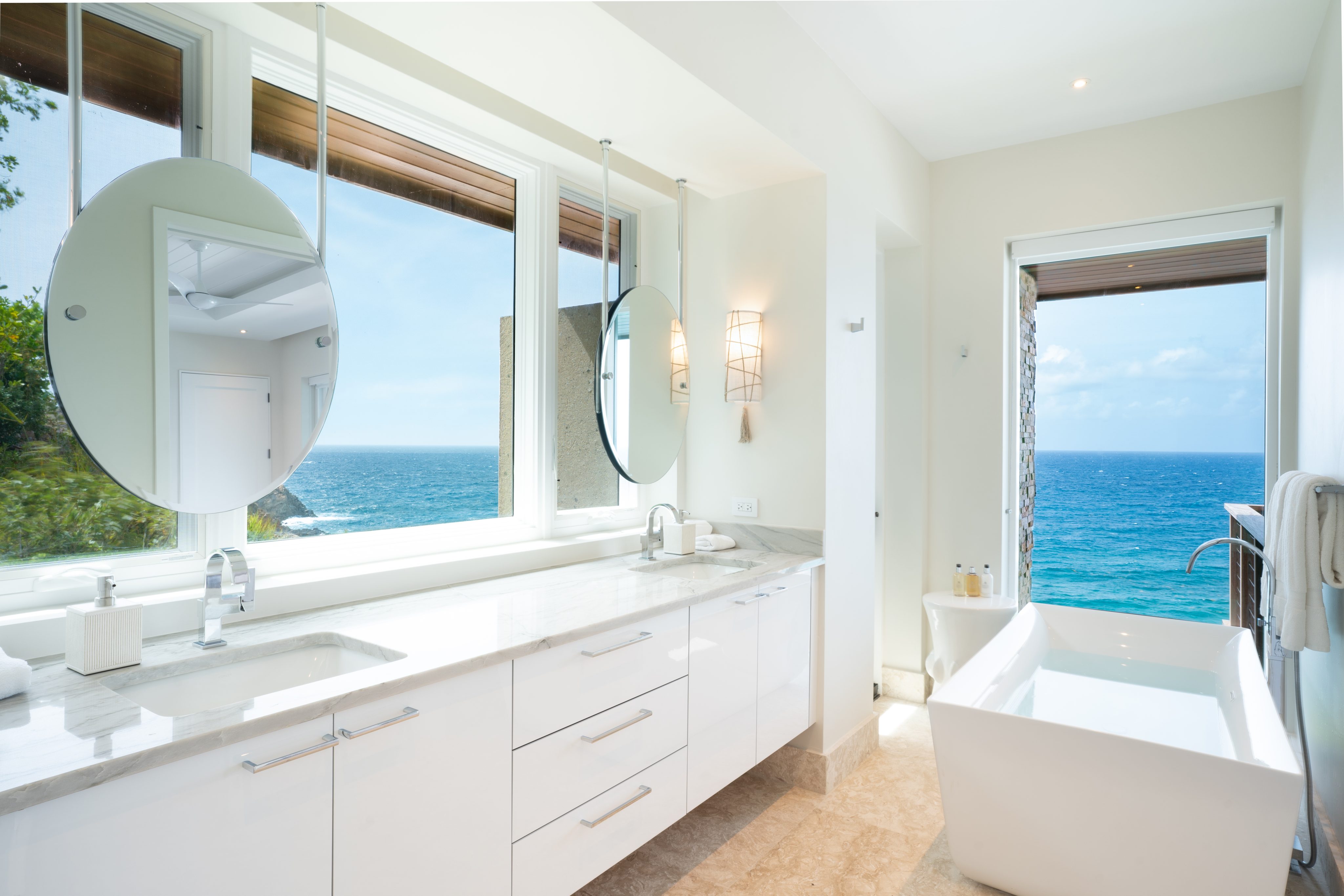 A bright, modern bathroom at Whale Point Villa features white cabinets, a double sink, and large circular mirrors. A freestanding bathtub is positioned near a window and glass door, offering a stunning ocean view. Natural light floods the space, highlighting the clean, minimalist design.