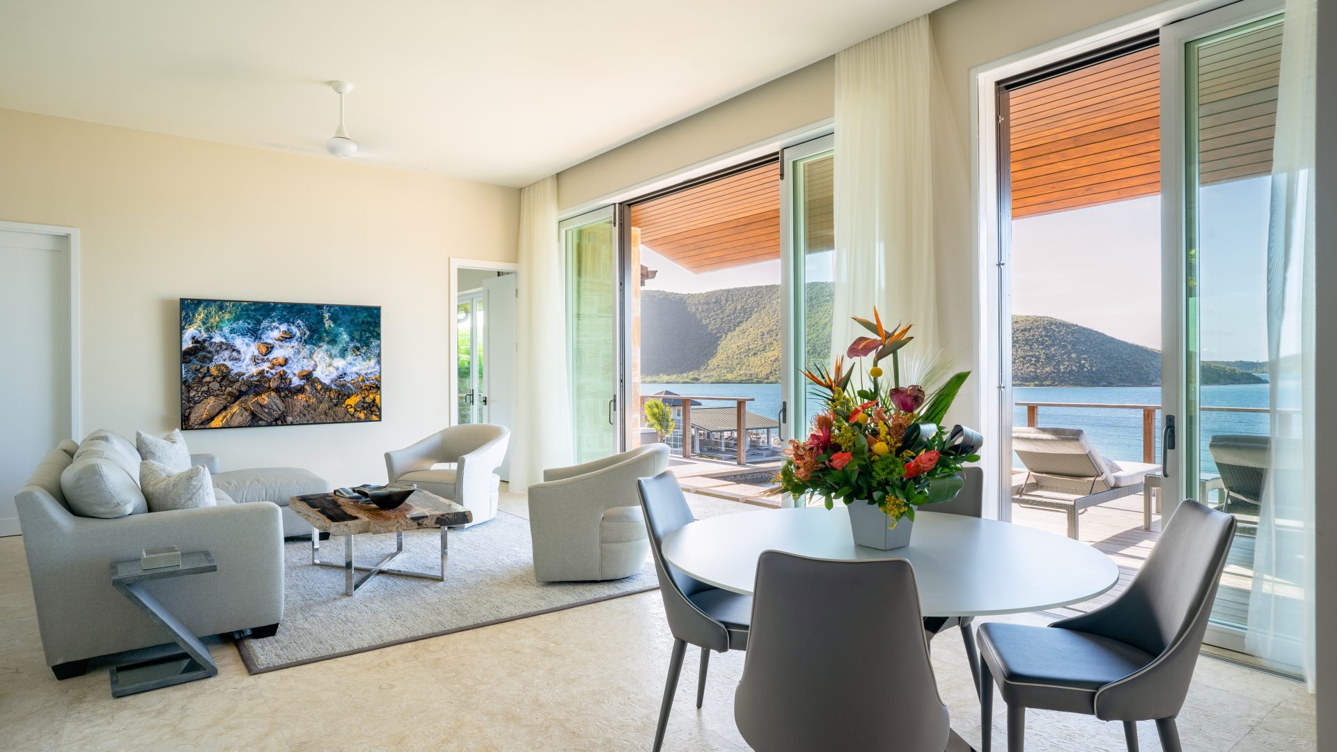 A bright and modern living room at Winedown Villa boasts large windows overlooking a scenic lake and mountains. The room features a plush sofa, two armchairs, a coffee table, and a dining table with four chairs. A colorful flower arrangement sits on the dining table, and a painting graces the wall.