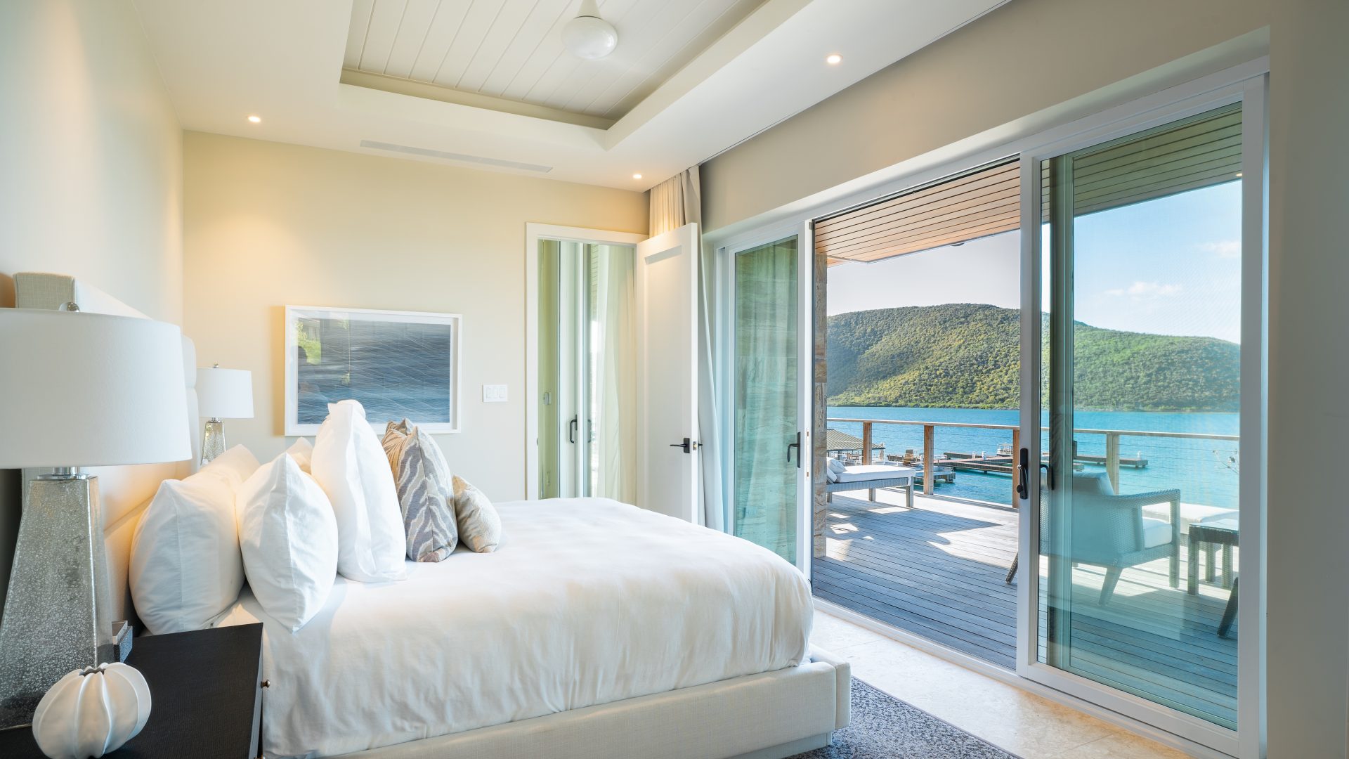 A modern, minimalist bedroom at Winedown Villa features a large white bed, neutral walls, and a sliding glass door leading to a balcony overlooking a scenic body of water and green hills. The room is bright and airy, with natural light streaming in through the door.