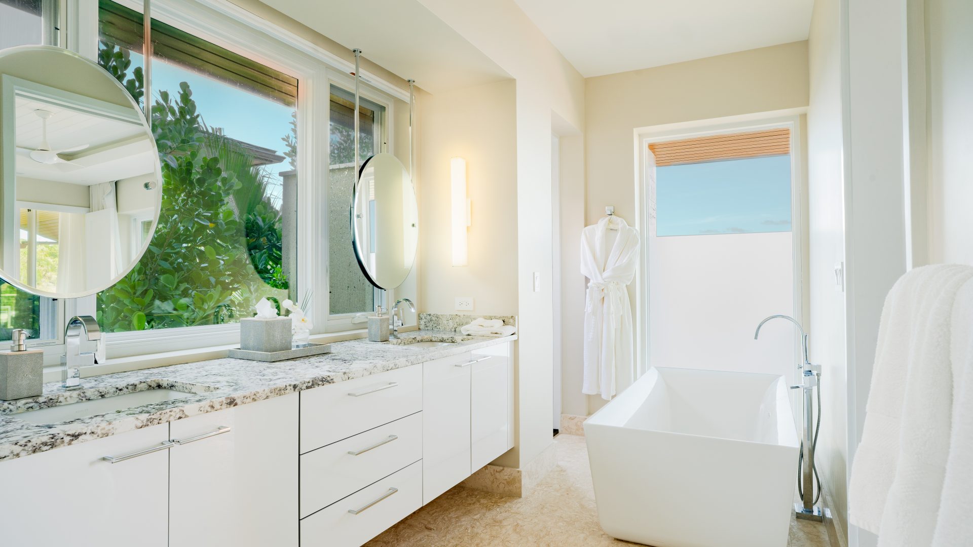 A modern bathroom at Winedown Villa features large windows, a marble countertop with double sinks, a circular mirror, a white freestanding bathtub, and a towel hung on the wall. A white robe hangs on the door, and lush greenery can be seen outside the window.