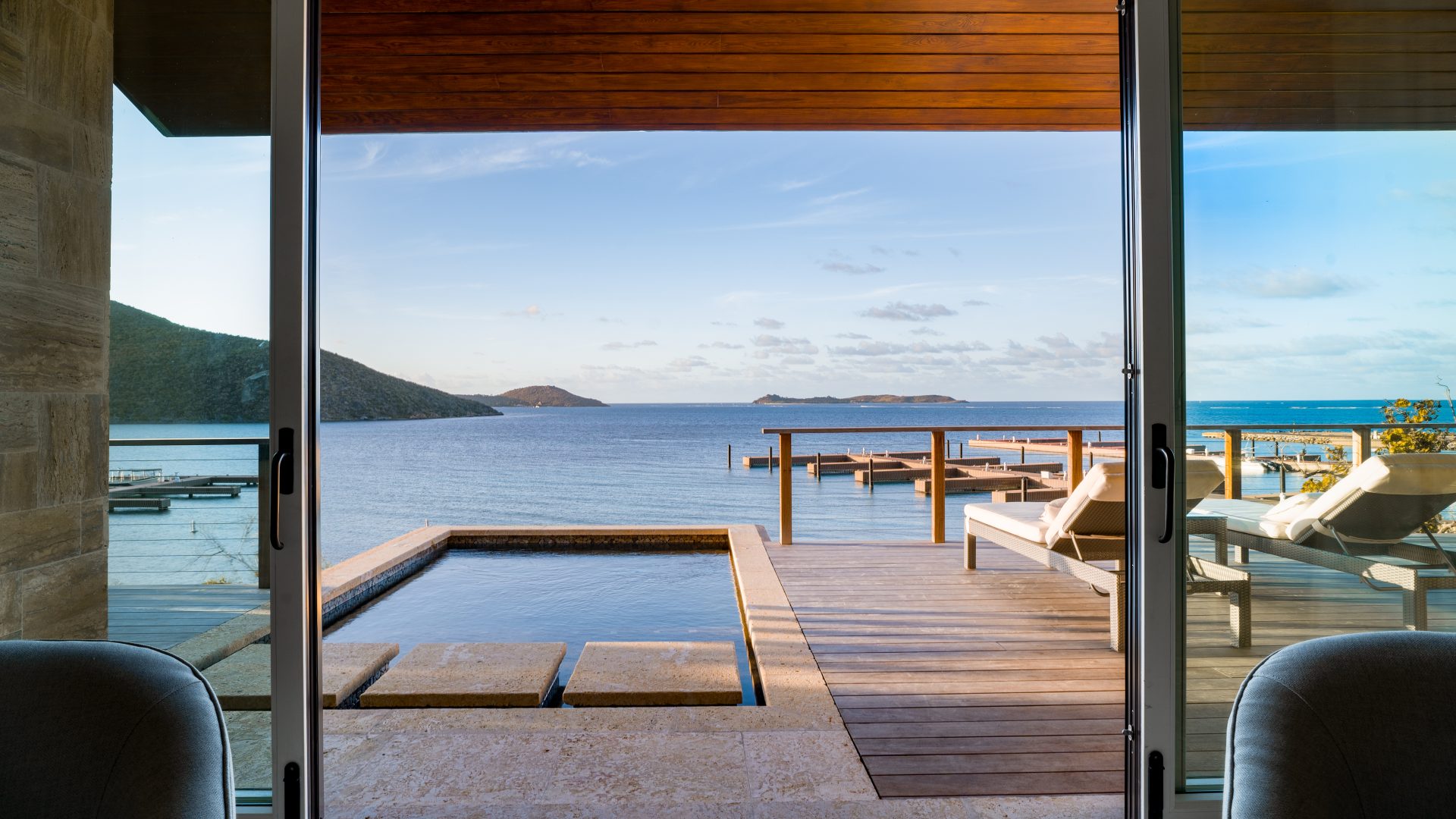 A view through a large glass door reveals the villa's luxurious patio with lounge chairs, an infinity pool, and a serene ocean landscape. The setting includes mountains in the distance under a clear sky, creating a peaceful and inviting atmosphere perfect for a winedown.