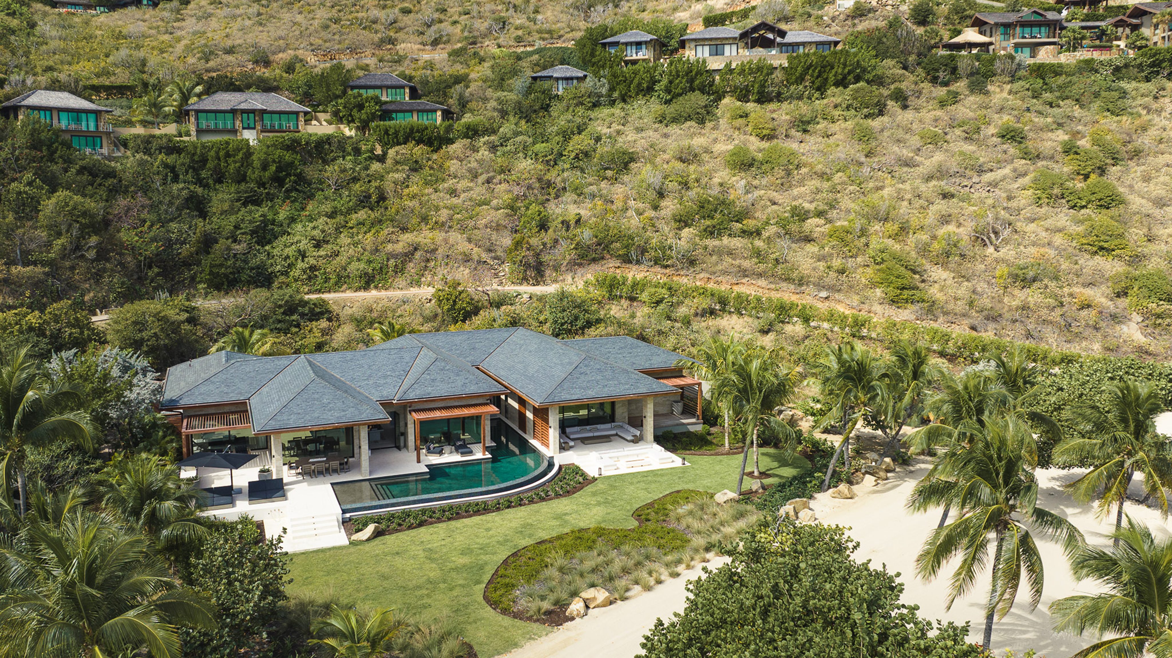 A luxurious beachfront villa, named Villa Xela, boasts a private pool surrounded by lush greenery and palm trees. The sleek modern architecture features multiple roof sections, large windows, and outdoor seating areas. Additional smaller villas are nestled on the hillside behind it.