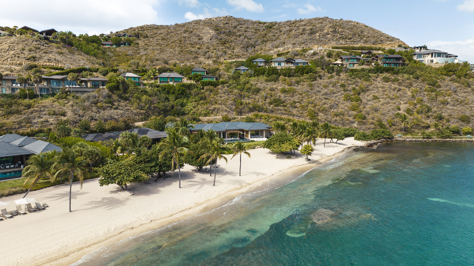 Aerial view of a pristine beach lined with lush palm trees and luxurious beachfront houses, including the stunning Xela Villa, on hilly terrain. Clear blue-green ocean water laps against the white sandy shore, with more residential structures nestled into the hilly landscape in the background.