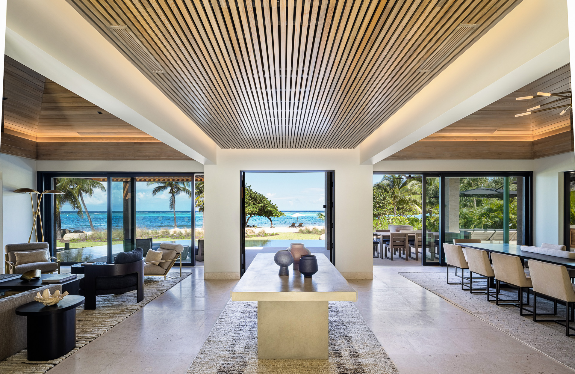 A modern beachfront villa living room with sliding glass doors that open to an ocean view. The room features a textured ceiling, neutral-toned furniture, and a long central table. Palm trees and blue skies are visible outside, creating a serene and inviting atmosphere.