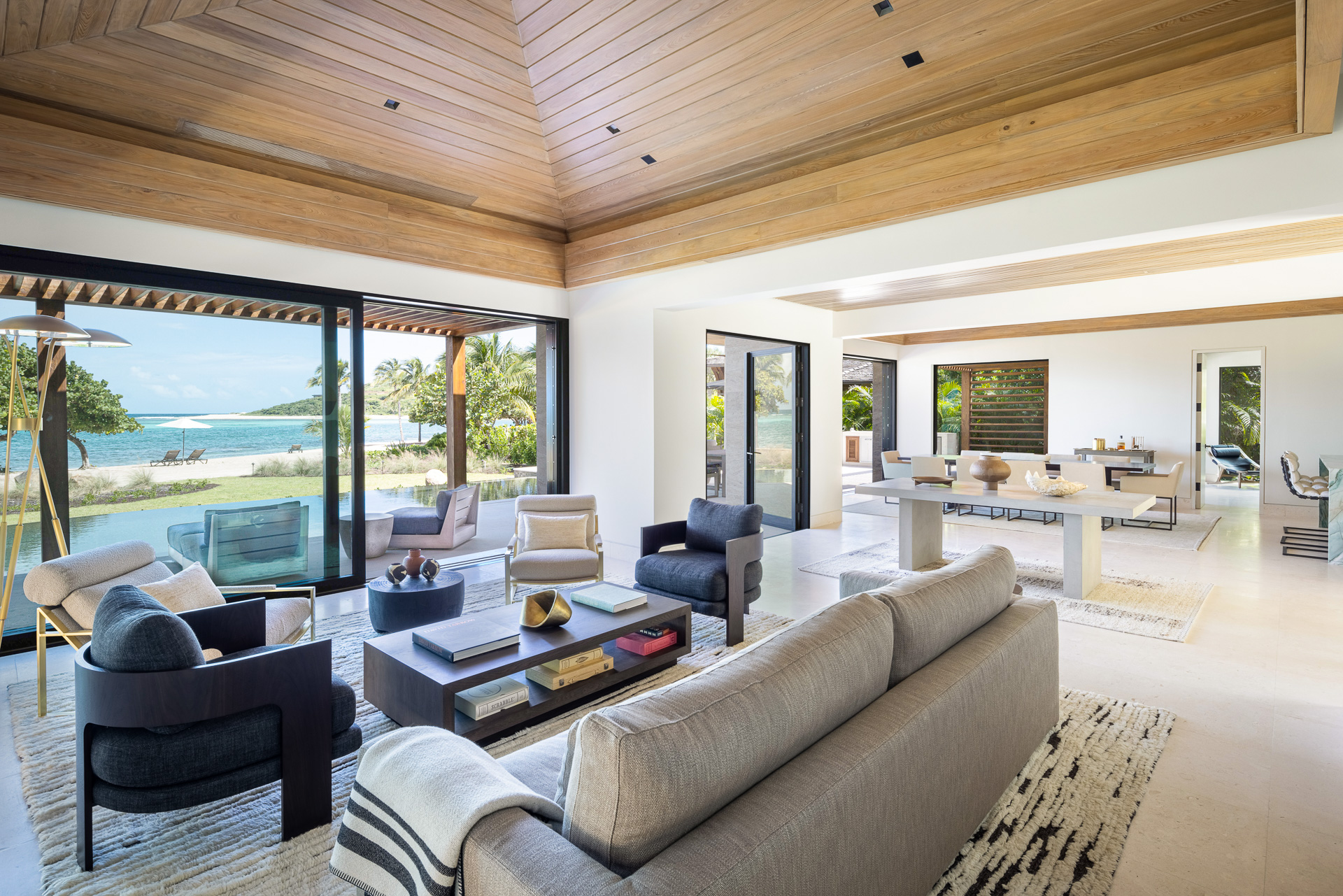 A spacious, modern living room in Villa Xela features a wooden ceiling and is furnished with a mix of light and dark contemporary furniture. Large sliding glass doors open to a scenic beach view, allowing natural light to flood the room. A dining area is visible in the background.