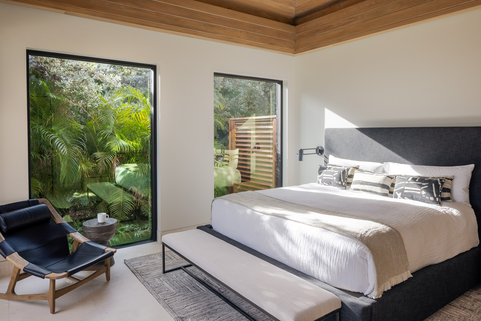 A modern bedroom in Villa Xela featuring a large bed with a dark headboard and white bedding, flanked by two windows showcasing lush greenery outside. A black lounge chair and wooden table are positioned in the corner. The room has light walls and a wooden ceiling, creating a serene atmosphere.