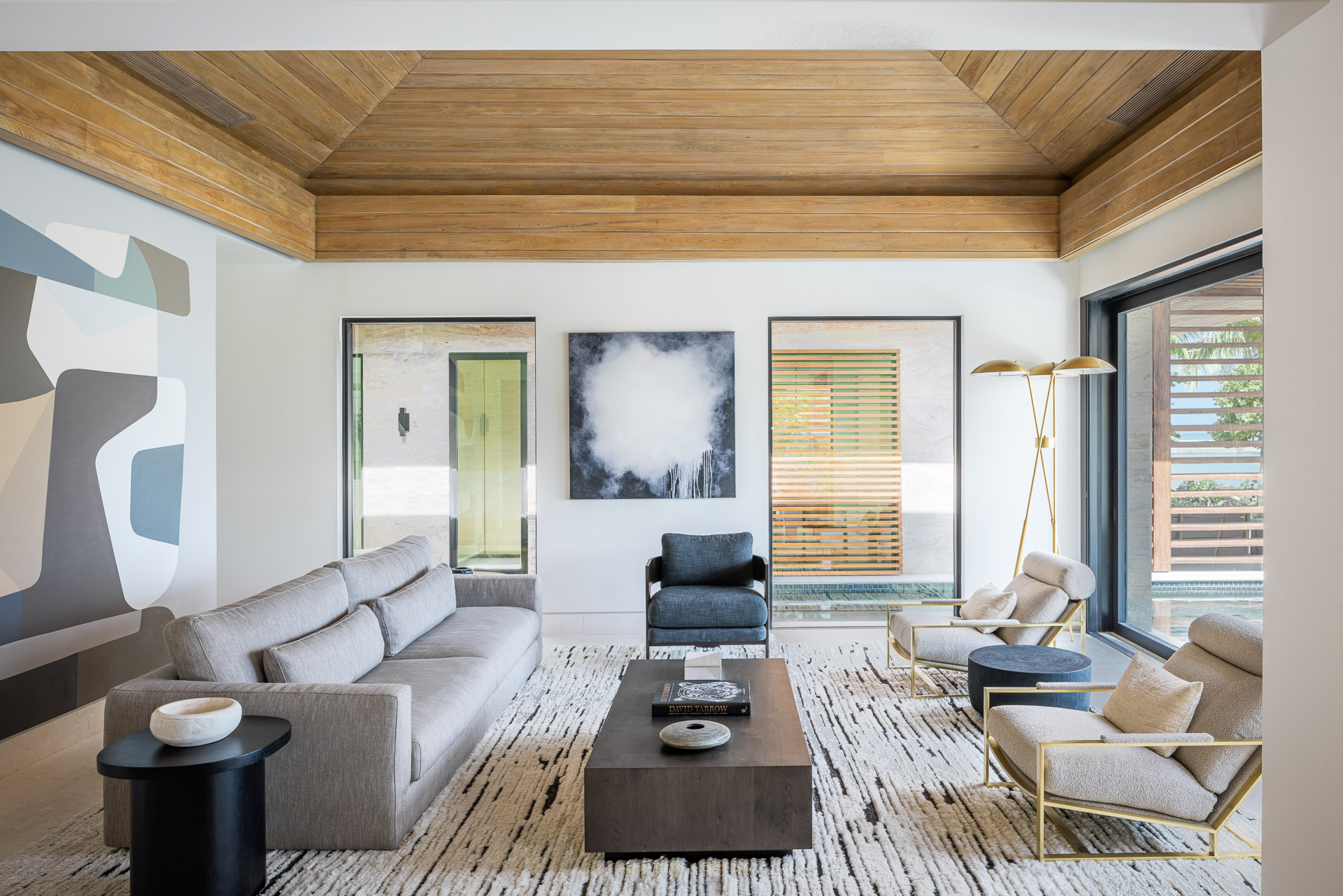 In Xela Villa's modern living room, a wooden vaulted ceiling crowns two large windows, a gray sofa, two cream armchairs with gold frames, a black armchair, and a black coffee table. A minimalist artwork graces the wall, complemented by a geometric mural on the left and a floor lamp on the right.