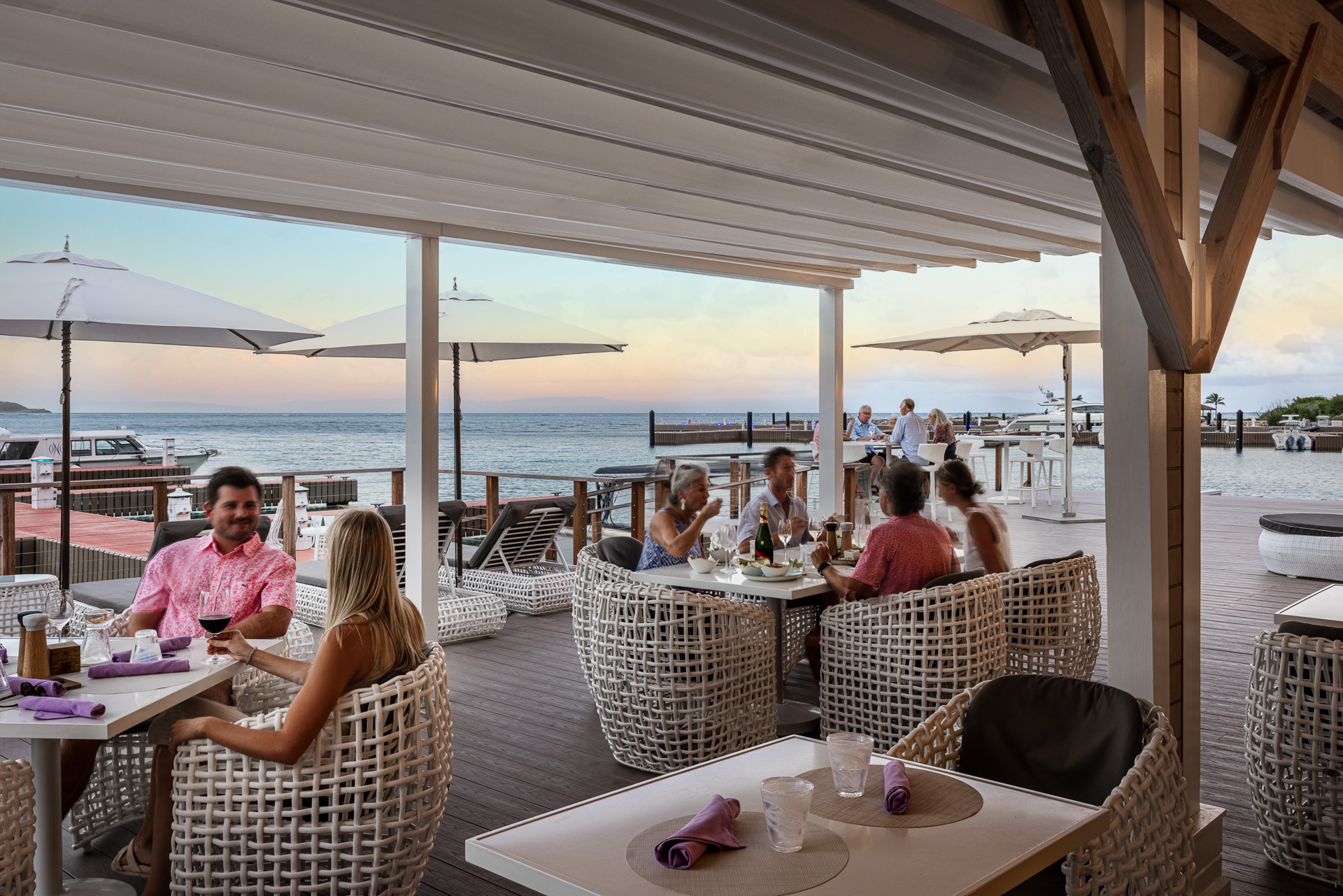 Guests at Nova lounge on wicker chairs under large white umbrellas, savoring drinks and conversation on a covered oceanfront patio. The relaxed atmosphere is perfectly complemented by the scenic sea view and clear sky.