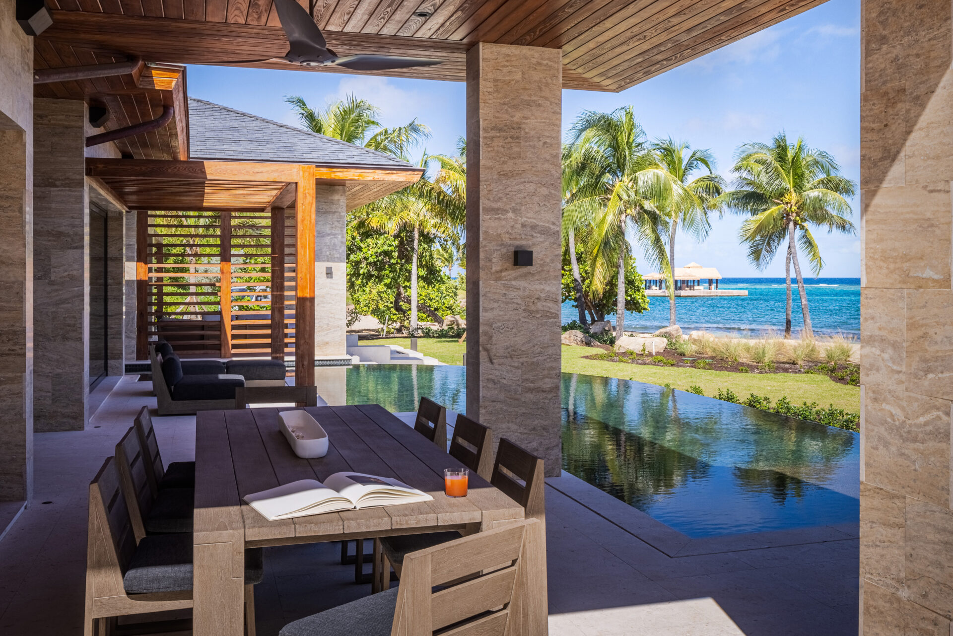 An outdoor patio at Villa Xela with a wooden dining table and chairs overlooks a sleek pool and a stunning beach scene with palm trees, a clear blue sky, and the ocean in the distance. An open book and a glass of juice sit invitingly on the table.