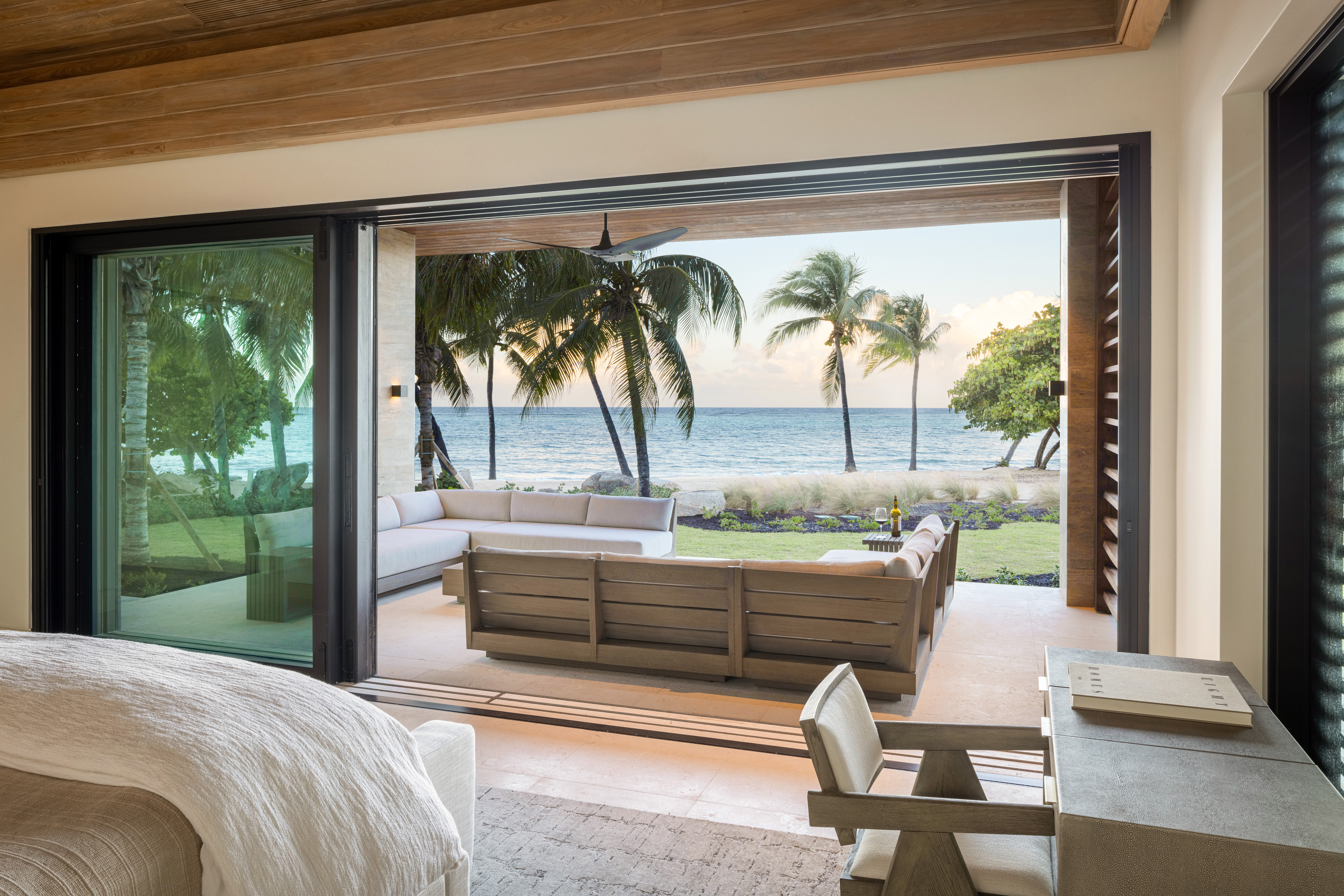 A modern bedroom at Xela Villa with sliding glass doors opens to a spacious patio featuring comfortable outdoor seating. The view includes palm trees, a grassy area, and a sandy beach with the ocean in the background. The room has a light, airy decor with neutral tones.