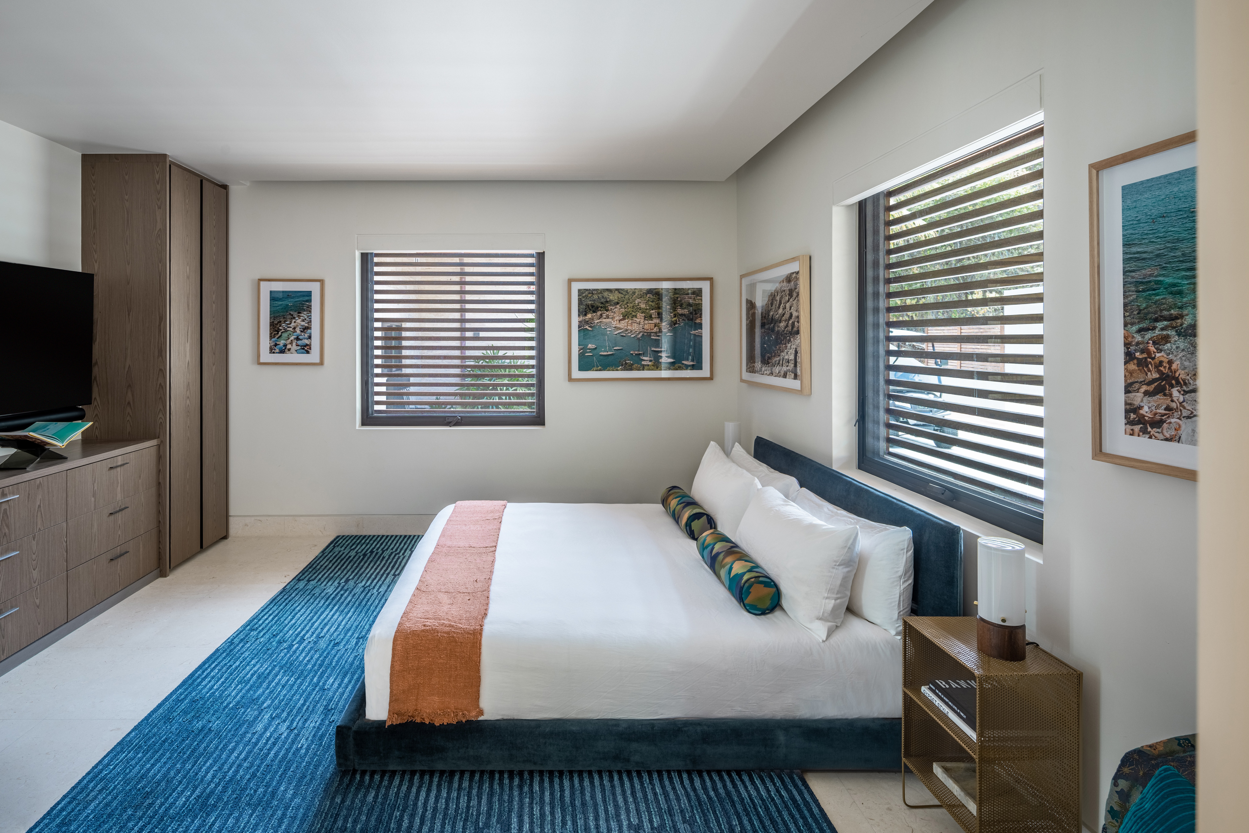 A modern bedroom in Xela Villa features a large bed with white and orange linens. The room includes a blue rug, light gray walls adorned with framed pictures, a window with horizontal blinds, a wooden dresser with a flat-screen TV, and a nightstand with a lamp.