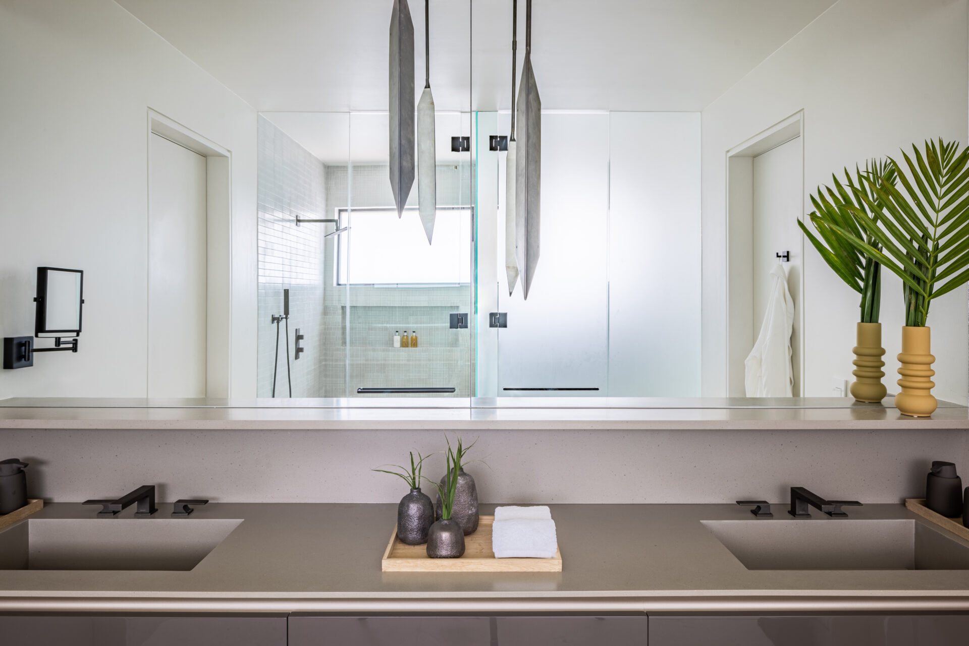 Modern bathroom in a Xela villa with a sleek double sink vanity featuring black fixtures and a tray with small plants and a folded white towel. In the background, there is a glass-enclosed shower with a window and toiletries. Large pendant lights hang from the ceiling, and a plant decorates the counter.