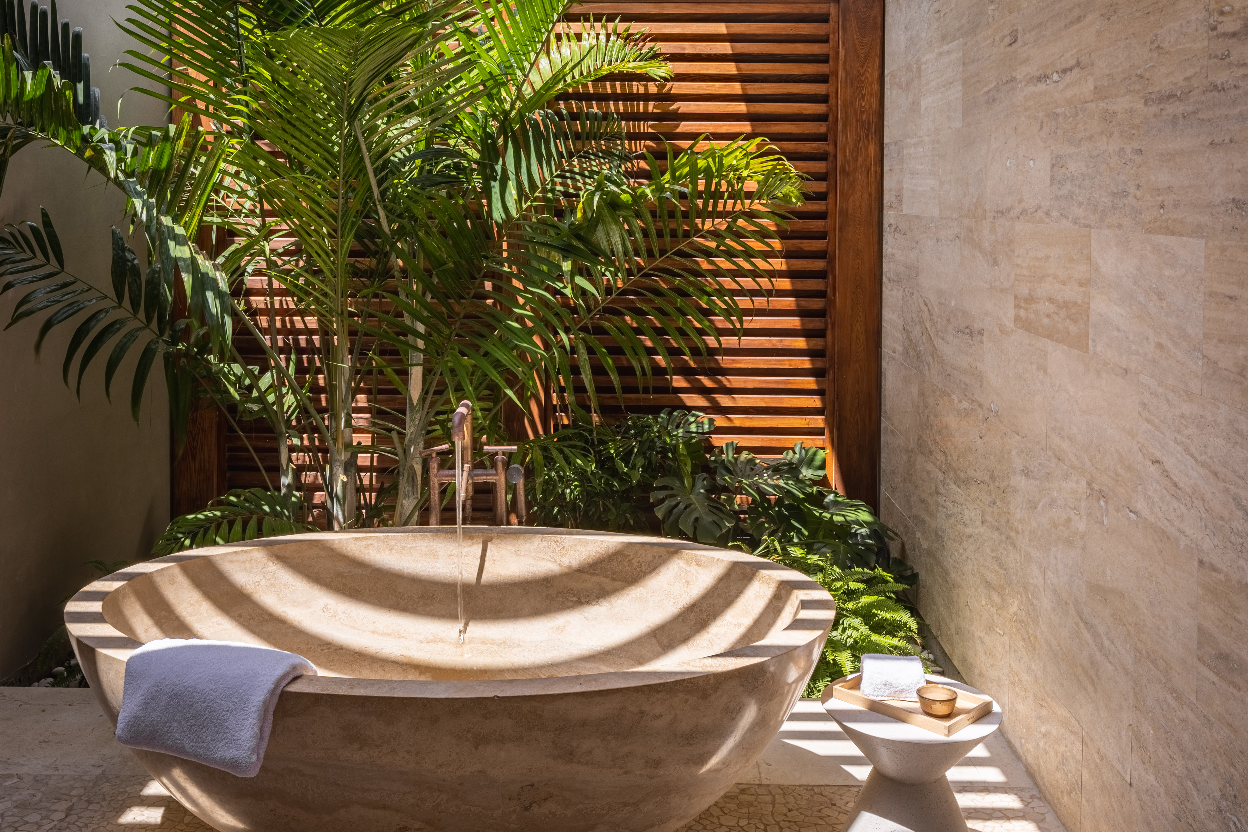 An outdoor bathroom in Villa Xela features a large stone bathtub surrounded by lush green plants. Sunlight filters through wooden slats, creating a serene and relaxing atmosphere. A small side table holds bath items, and a towel is draped over the edge of the tub.