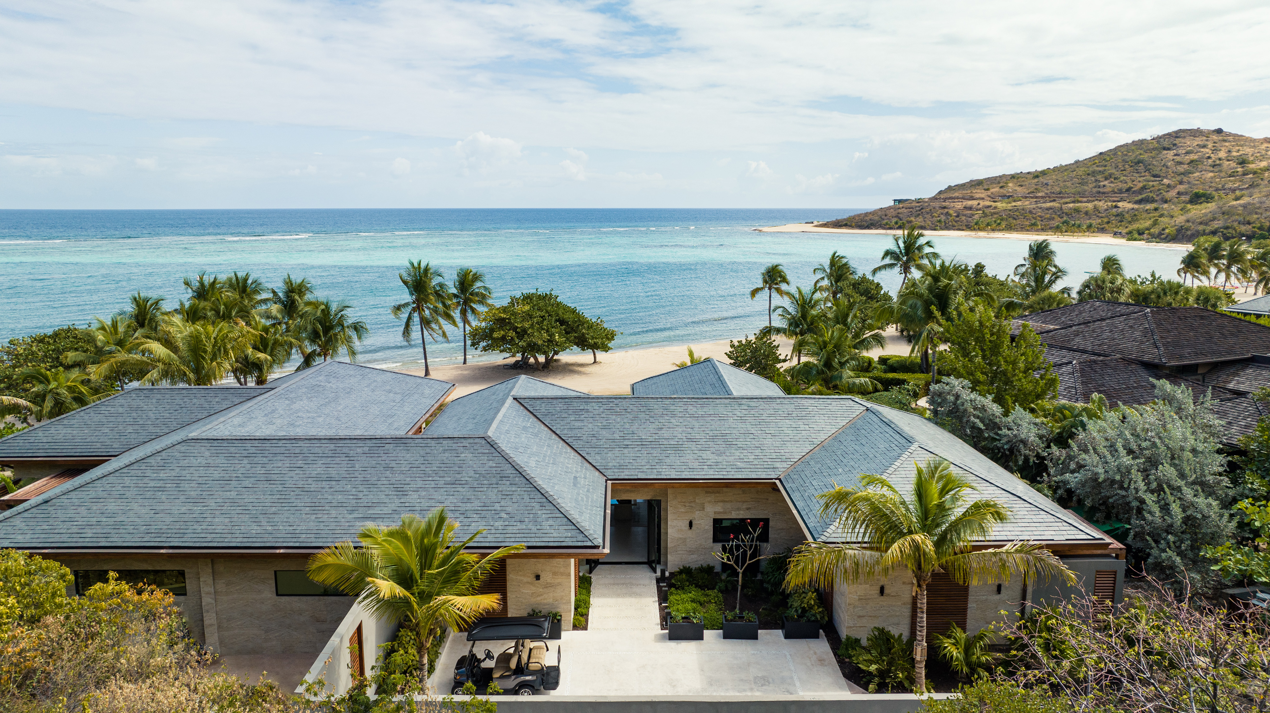 Aerial view of the luxurious Xela beachfront villa with a grey roof, surrounded by palm trees. The villa overlooks a serene, clear blue ocean, with a sandy beach and a distant hilly landscape in the background under a partly cloudy sky.