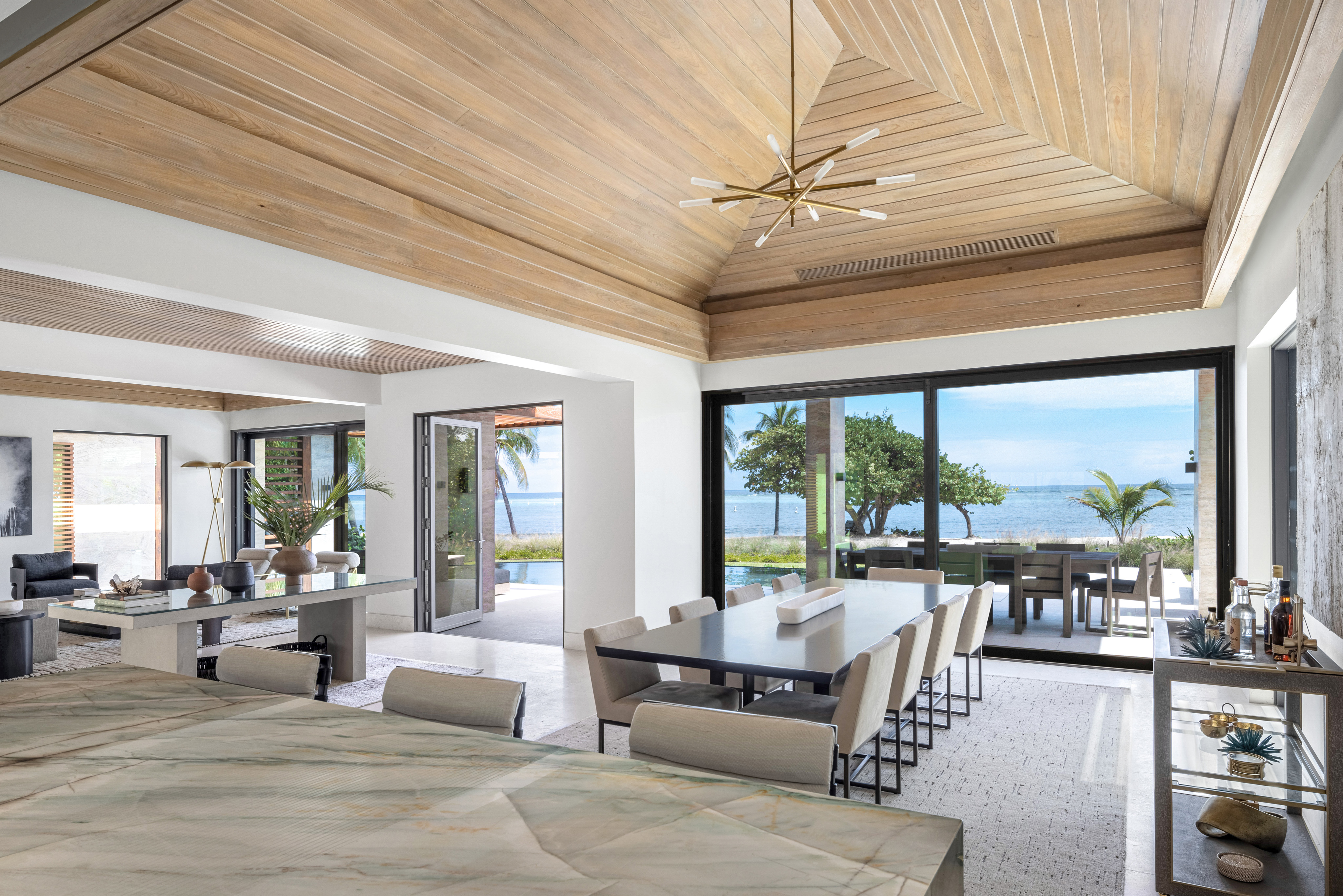 A contemporary open-plan living and dining area with a coastal view. The space in this Xela Villa features wooden ceilings and large windows, offering a scenic ocean view. Modern furniture fills the room, including a long dining table, a bar area, and a partially visible marble counter.