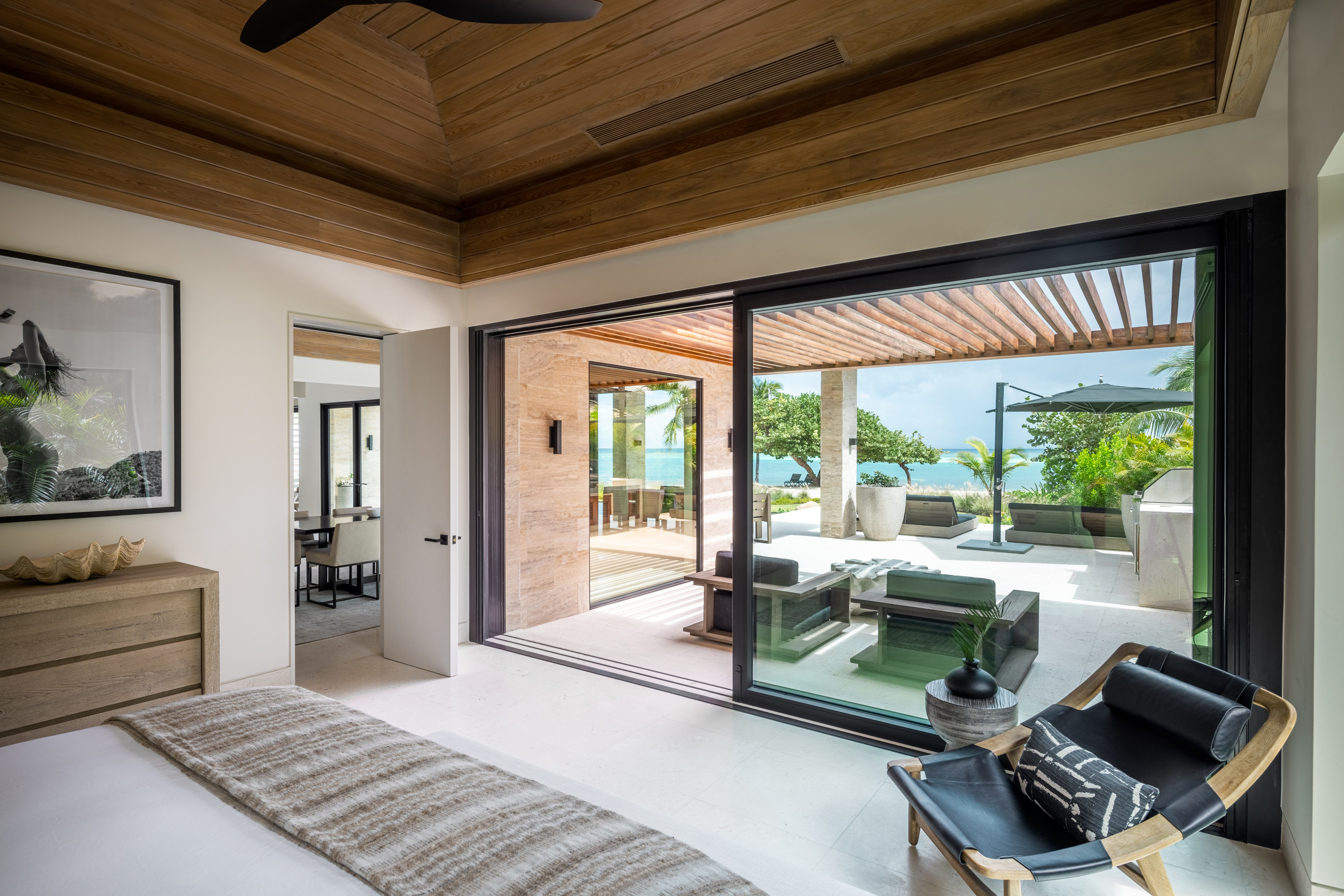 A modern, spacious bedroom with a wooden ceiling and light-colored floor opens to an outdoor patio through large glass sliding doors. The Xela Villa patio features a seating area, lush greenery, and a view of the ocean under a partially covered pergola.