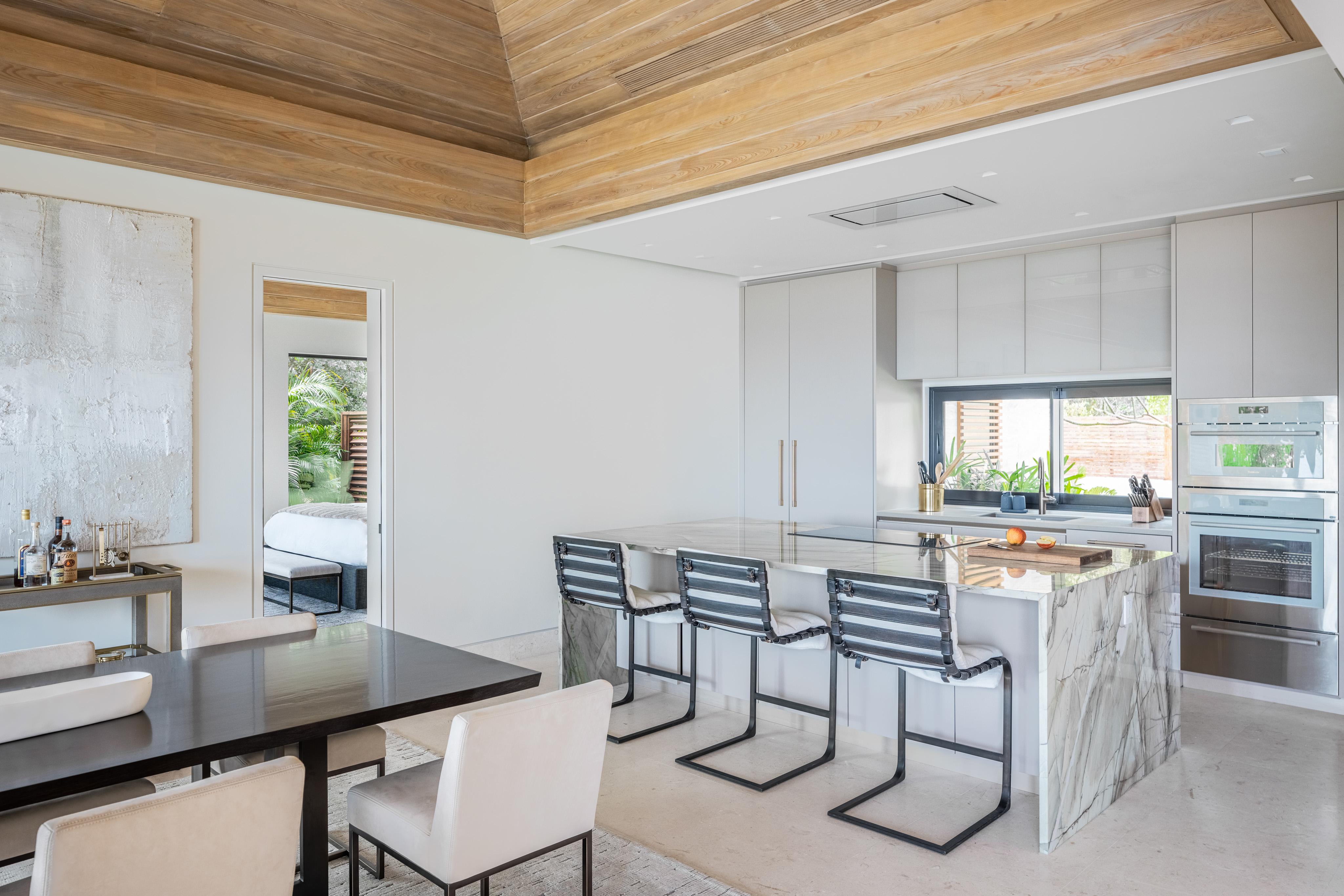 Modern open-plan kitchen and dining area in Villa Xela with a high wooden ceiling. The kitchen features a marble island with four striped barstools, stainless steel appliances, and light gray cabinetry. A dining table with white cushioned chairs is in the foreground, and large windows let in natural light.