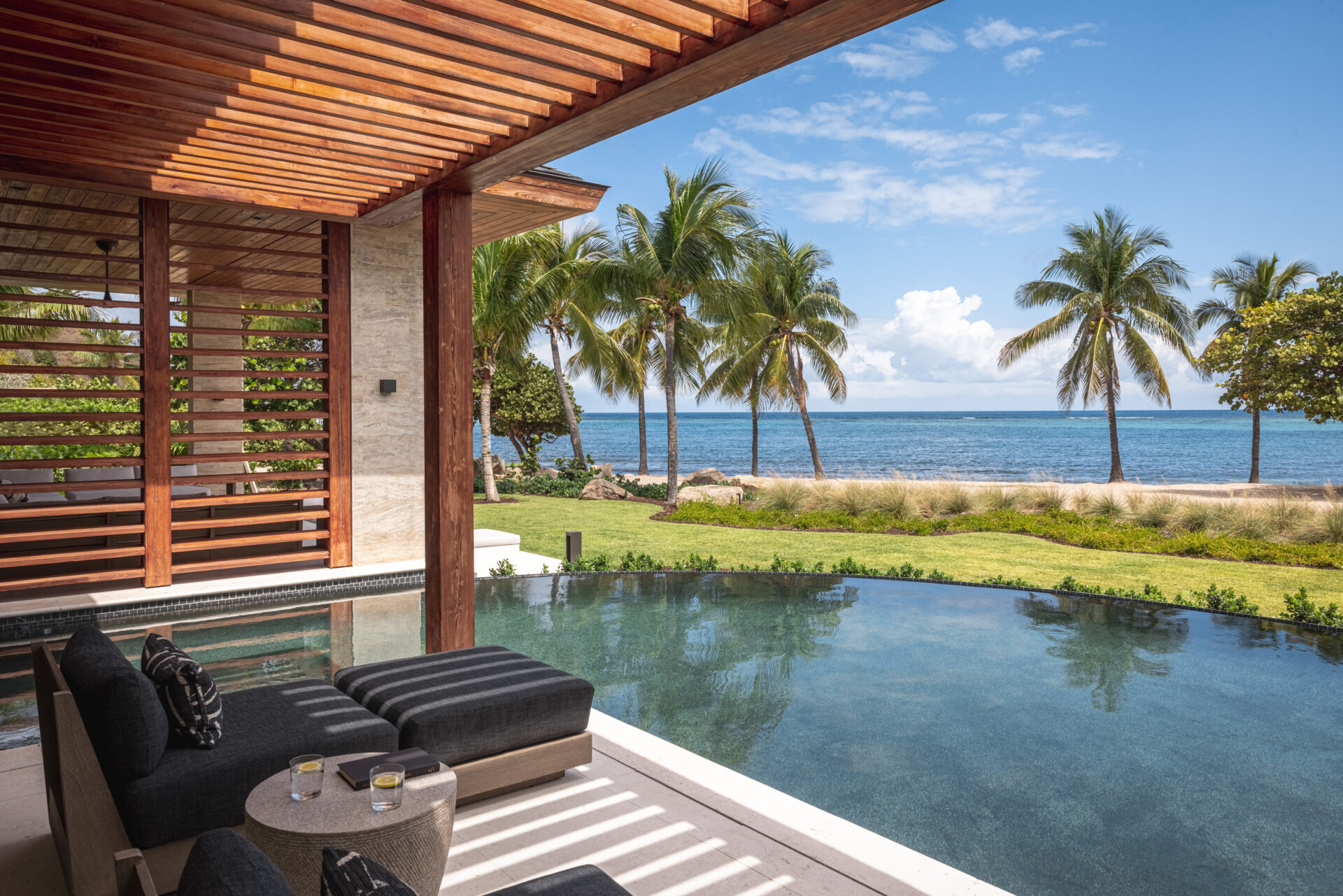 A luxurious outdoor seating area at Villa Xela features black cushioned furniture overlooking an infinity pool, palm trees, and a serene ocean view. The shaded wooden pergola provides a perfect blend of sunlight and shadow, creating a calm and tropical ambiance.