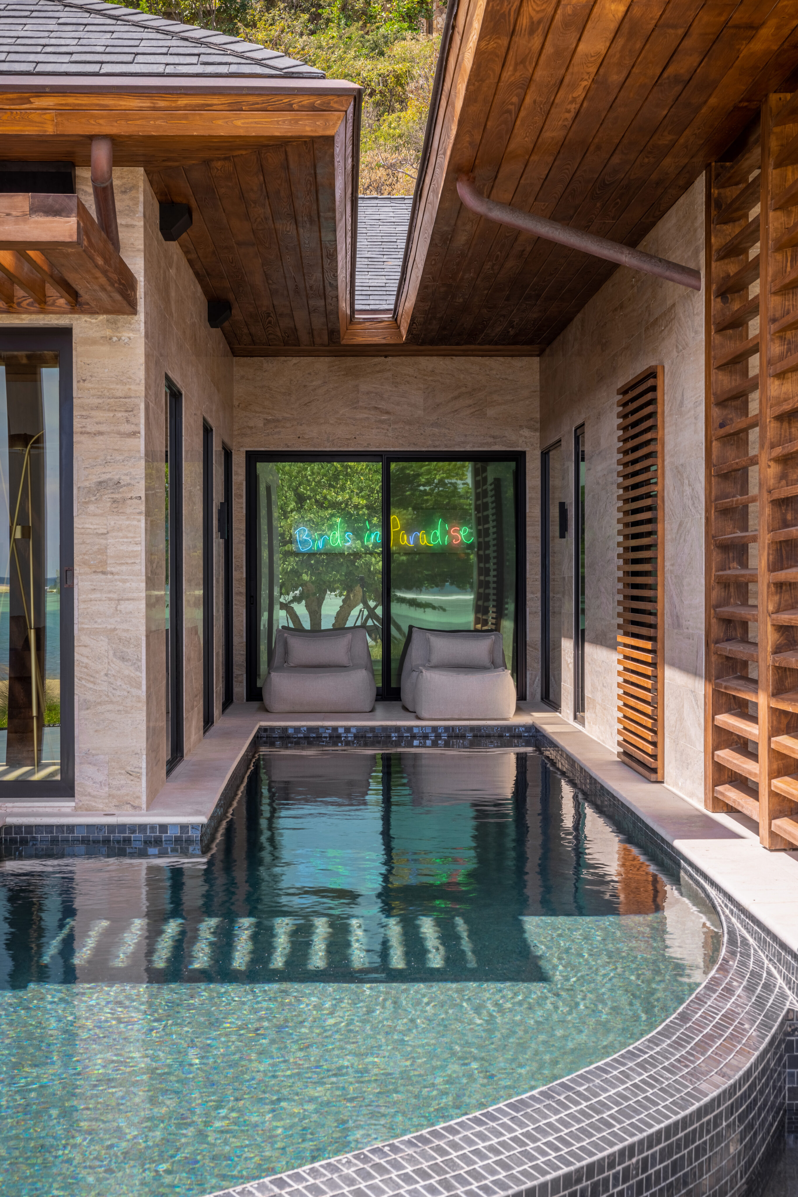 A serene, narrow pool is surrounded by a modern villa with wood accents and beige stone walls. Two lounge chairs sit by a glass door entrance adorned with a neon sign reading "Birds in Paradise." Lush greenery can be seen through the windows of this exquisite Xela retreat.