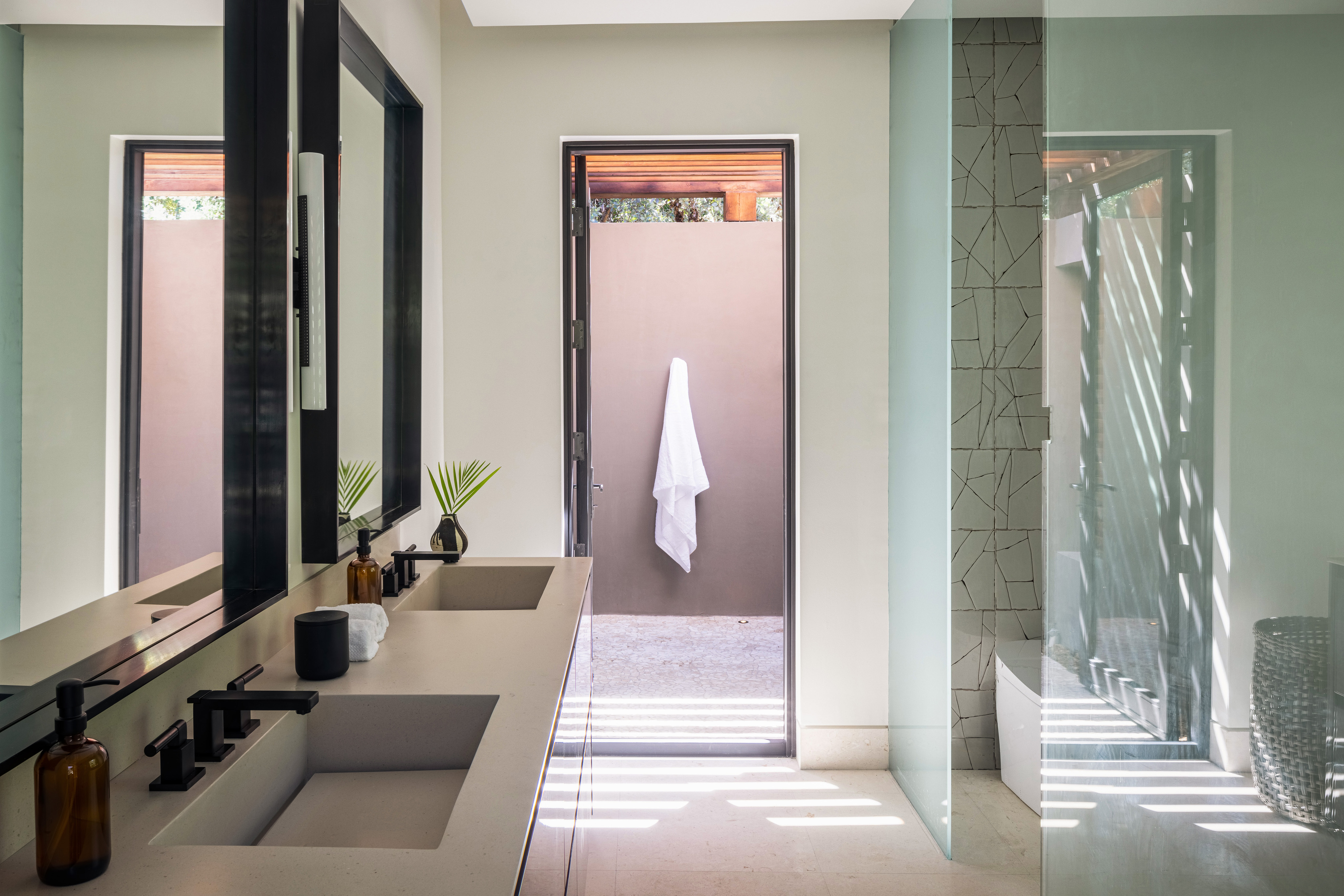 Modern bathroom in Villa Xela with a double sink vanity on the left, featuring black fixtures and decor. A glass-enclosed shower is on the right, showcasing a geometric tile design. An open door at the end of the room leads to a small patio with a towel hanging on a hook.