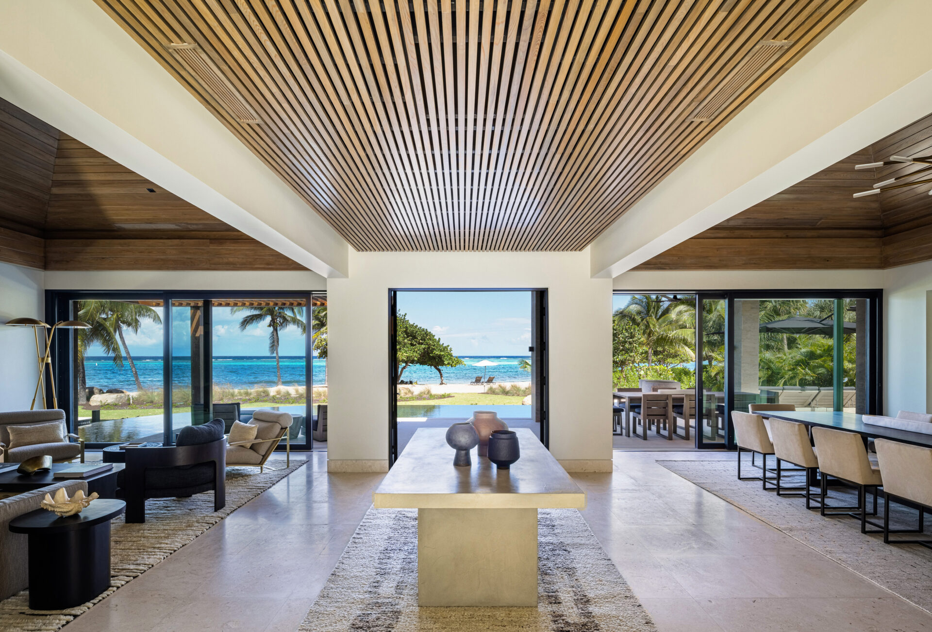 A spacious modern living and dining area with a high, wooden slatted ceiling awaits you at Xela Villa. Floor-to-ceiling sliding glass doors open to a view of the ocean. The room features minimalist furniture including a large central table, two sitting areas, and a dining set.