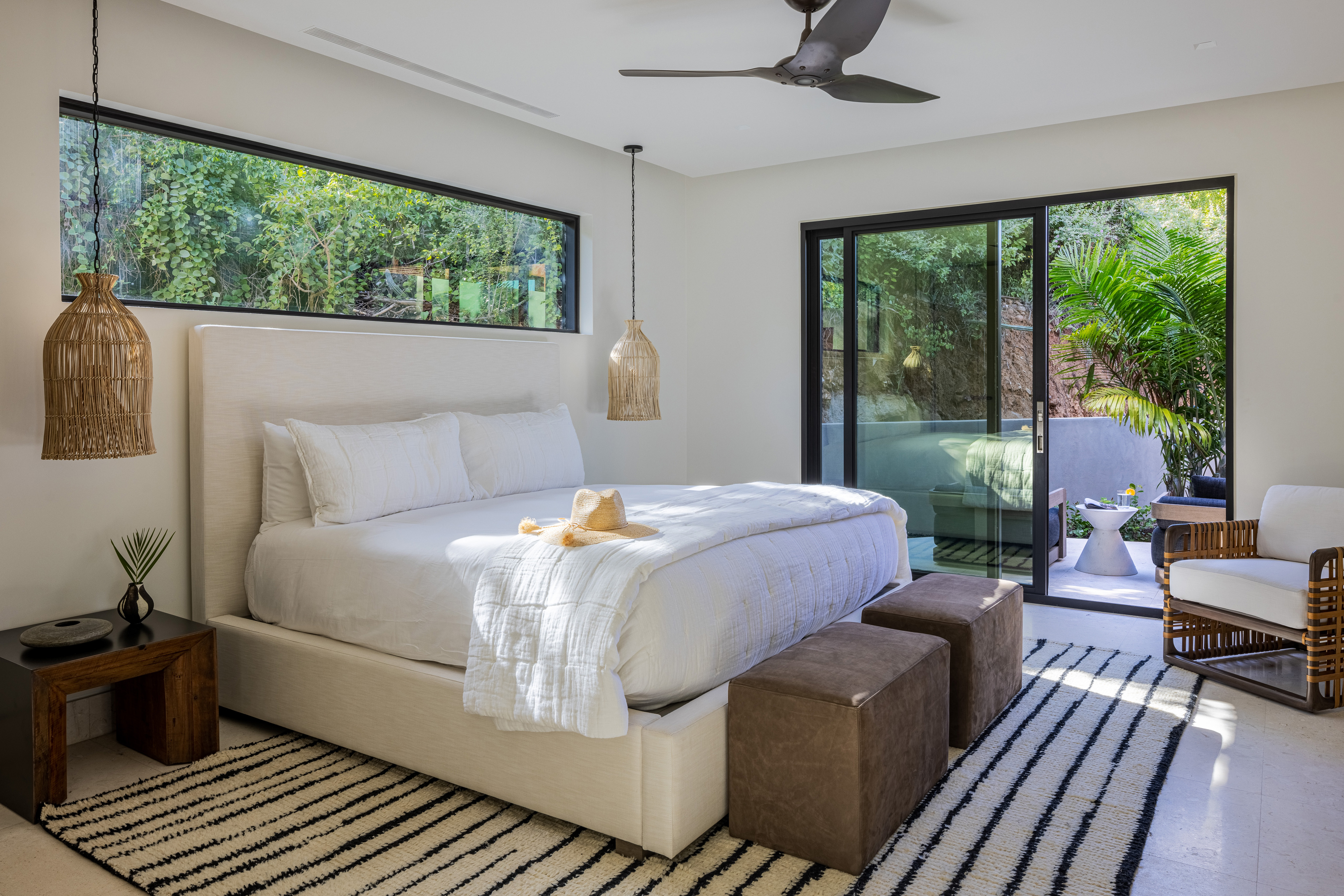 A bright bedroom in Villa Xela features a large bed with white linens and two brown ottomans at the foot. The room has hanging pendant lights, a striped rug, and large windows showcasing greenery outside. A modern ceiling fan above and a sliding glass door leads to an outdoor area.