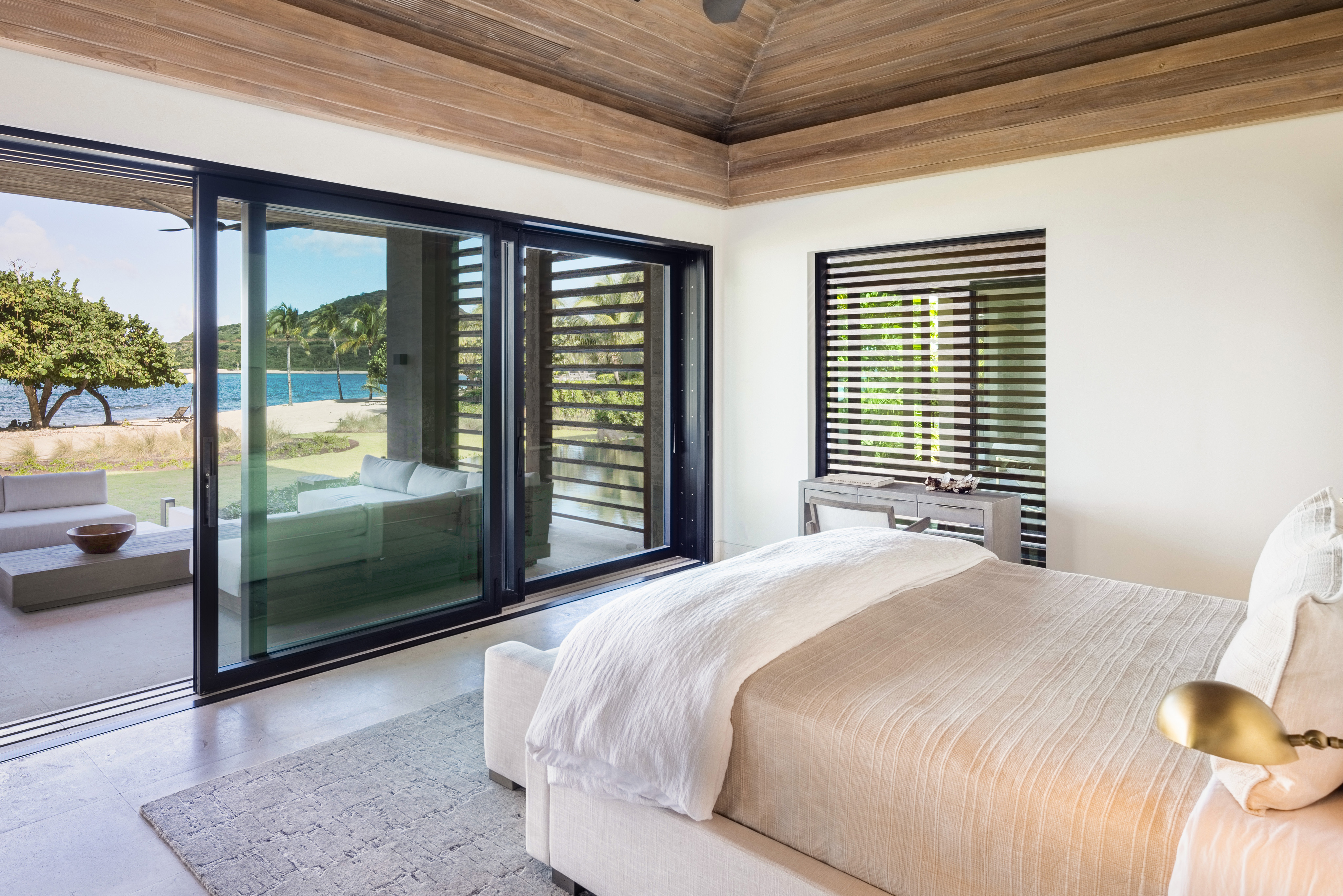 A modern, bright bedroom in Villa Xela features a large bed with white linens and cozy decor. Floor-to-ceiling sliding glass doors open to a terrace with lounge chairs, offering a view of a serene waterfront scene framed by palm trees. Natural light fills the room.