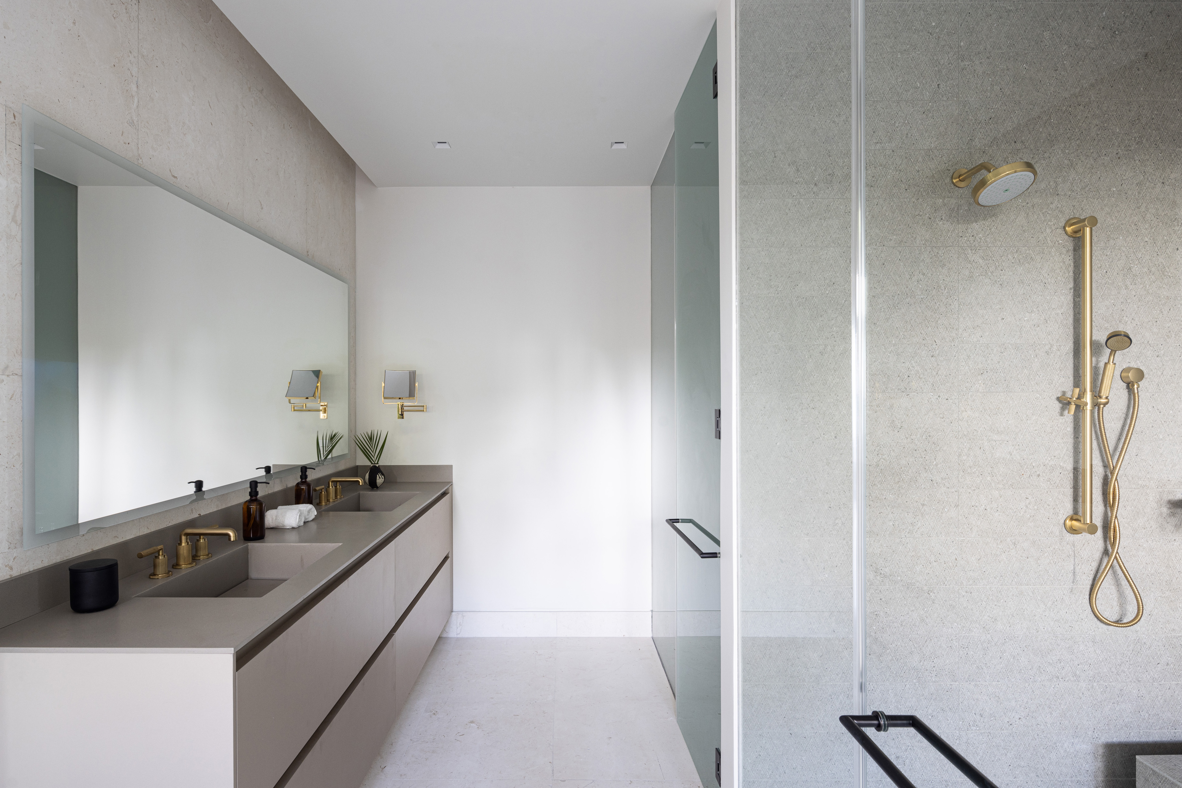 A modern bathroom in Villa Xela featuring a large mirror, double sink with sleek faucets, beige countertop, and cabinetry. On the right, there's a glass-enclosed shower with a rain showerhead and handheld shower. The room is lit with natural light, and the design is minimalistic.