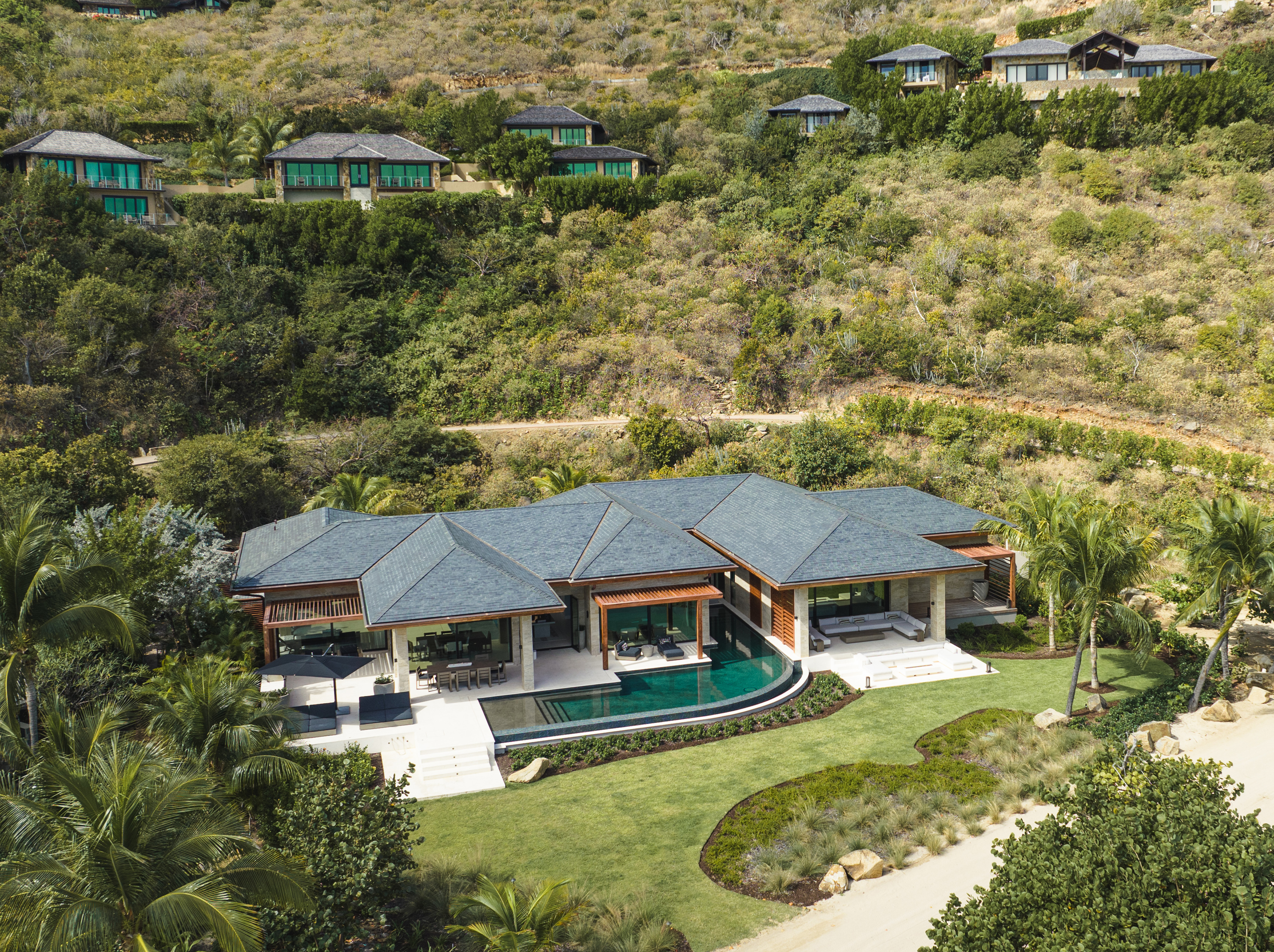 Aerial view of Xela, a luxurious hillside villa with a private pool, lush greenery, and multiple smaller buildings surrounding it. The modern architecture features large windows and a spacious outdoor area, nestled in a verdant landscape with a curving path nearby.