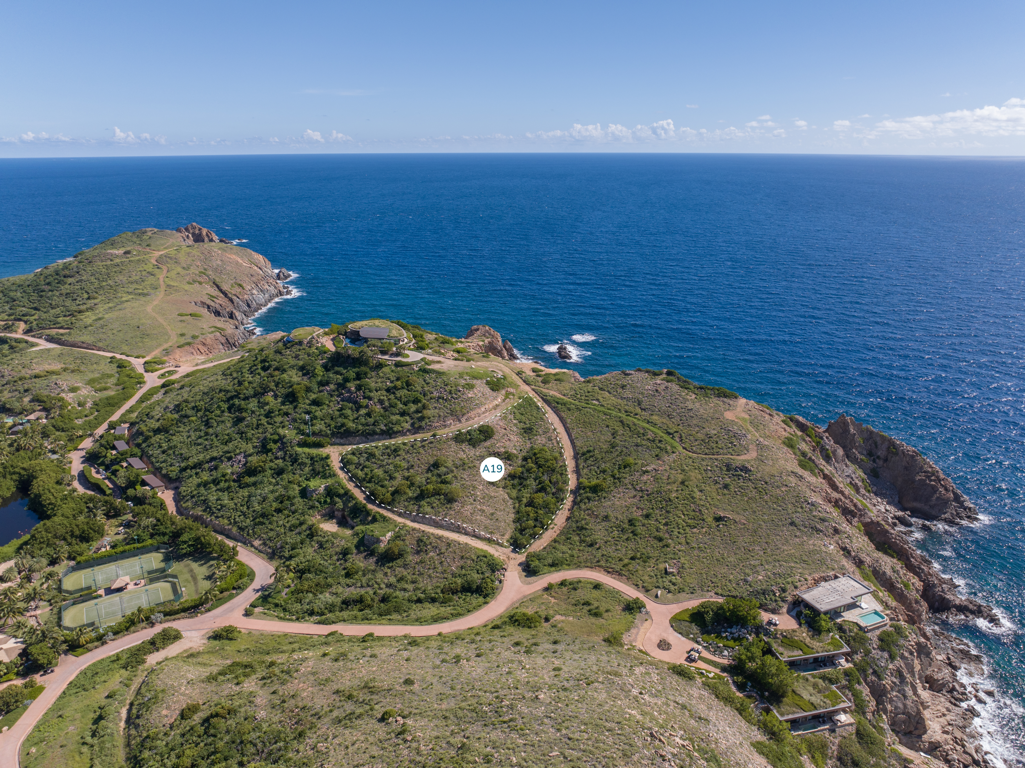 Aerial view of a coastal landscape featuring lush green hills, winding roads like the A19, and rugged cliffs meeting the blue ocean. Small homesites and structures are scattered across the area. The horizon is clear with a blue sky and a few scattered clouds over the Atlantic Ridge.